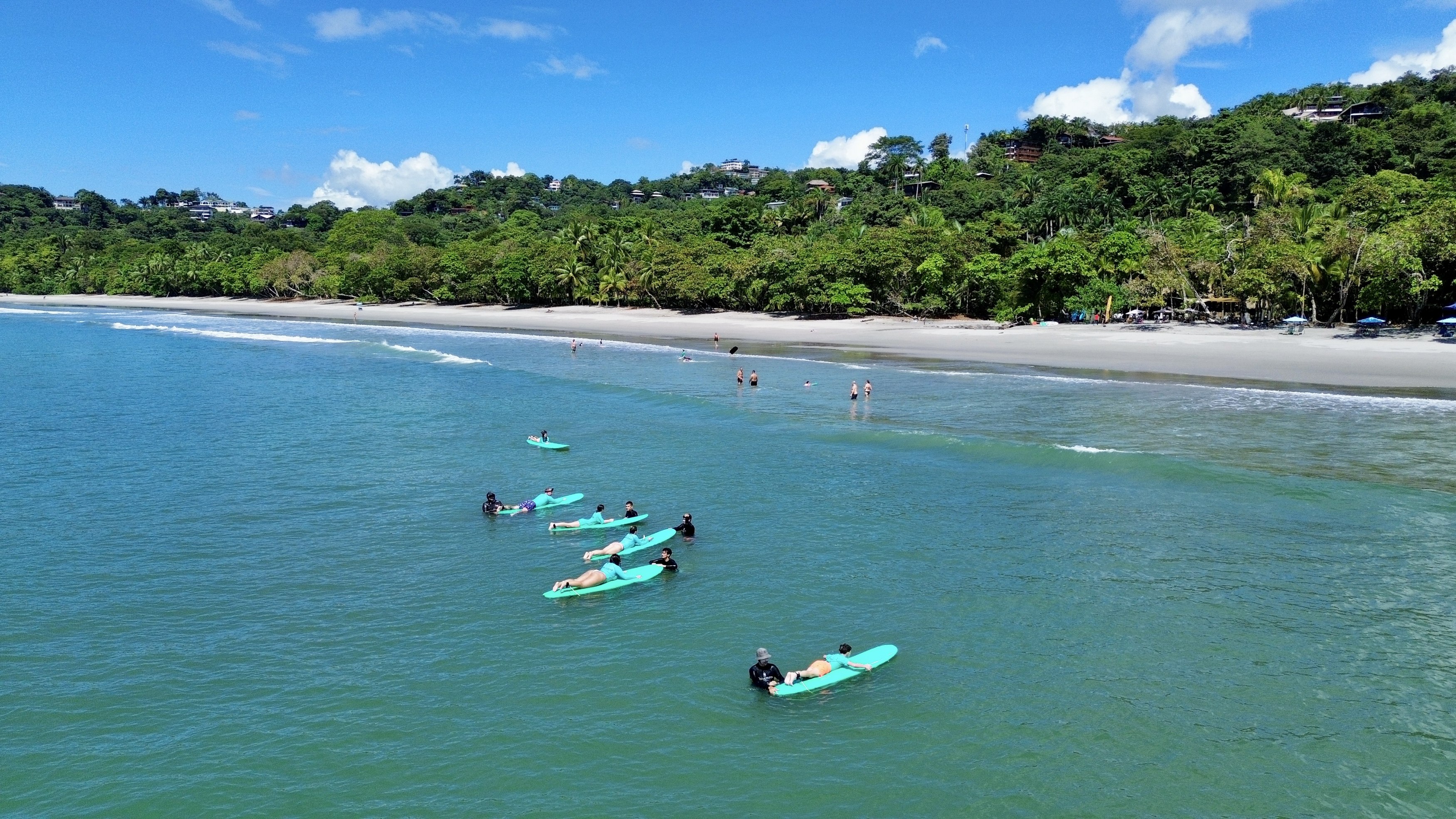 An aerial view shows clear turquoise ocean waters where people are swimming and learning to surf, leading to a sandy beach backed by a lush green hillside dotted with homes under a bright blue sky.