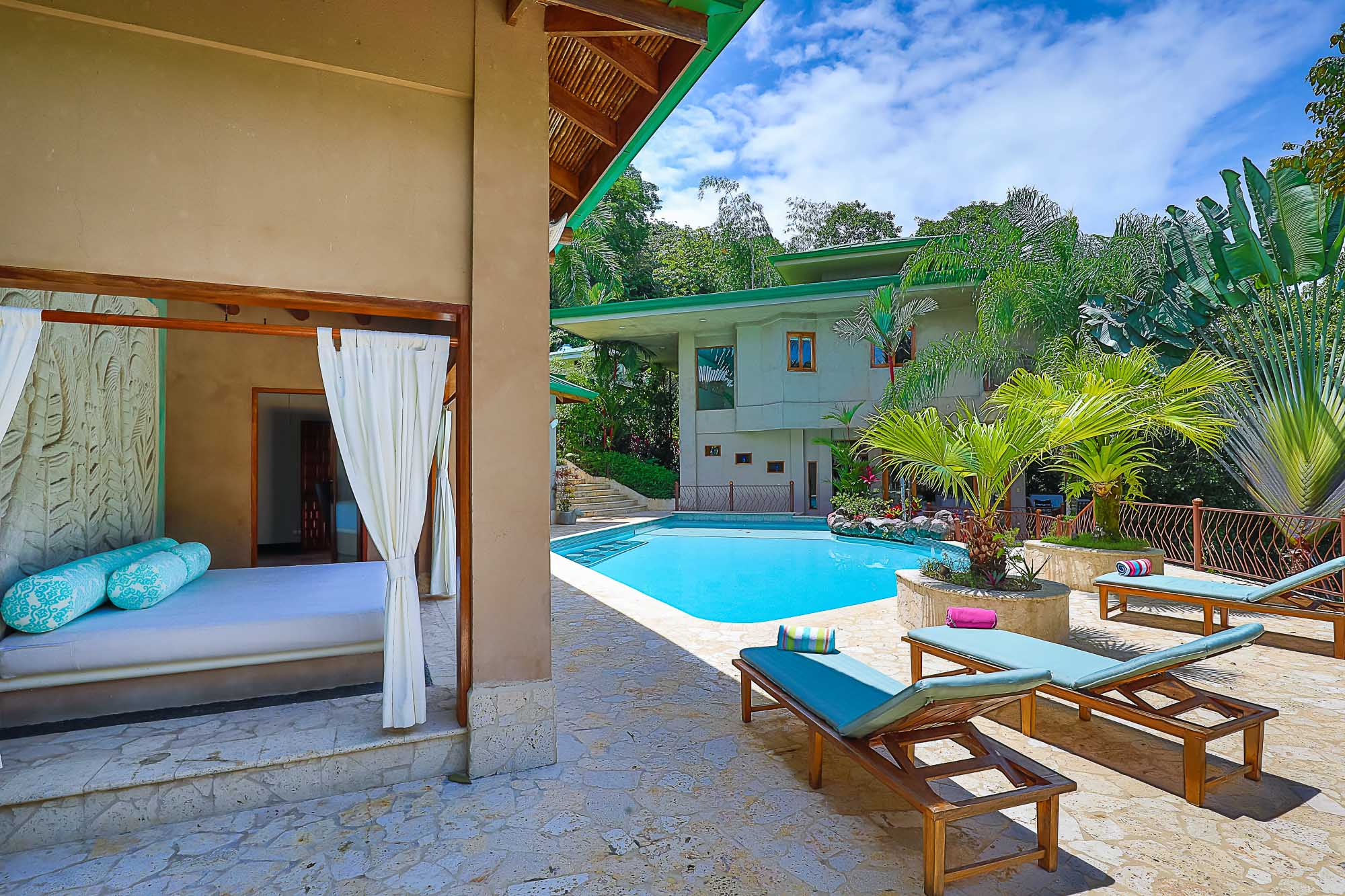 A bright outdoor area features a large swimming pool, sun loungers on a stone patio, and a shaded pavilion with a daybed, all set amidst tropical gardens with views of the main villa.