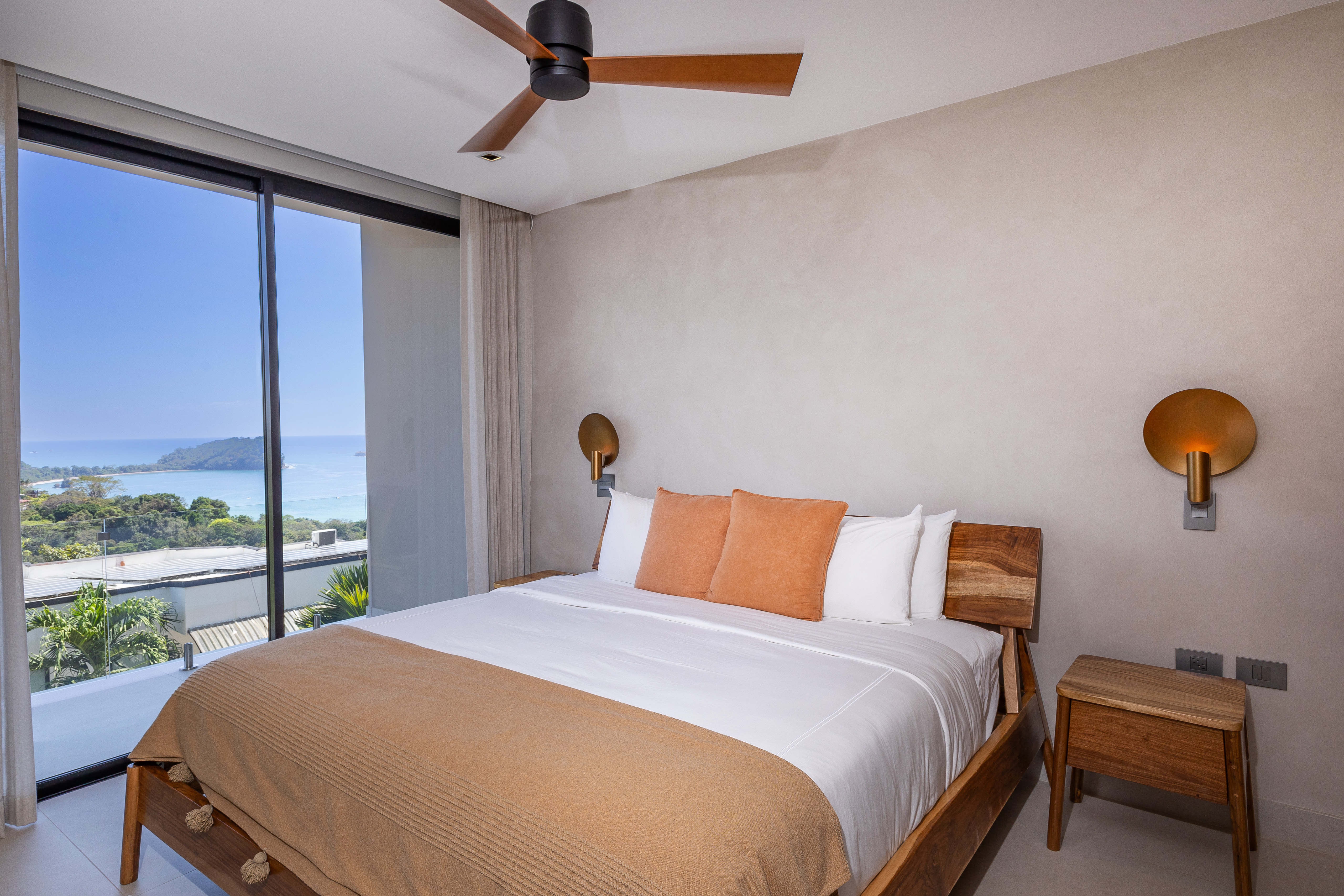 This bedroom offers a large bed, a modern ceiling fan, and full-height glass doors that open to a panoramic ocean and jungle view, filling the space with natural light.
