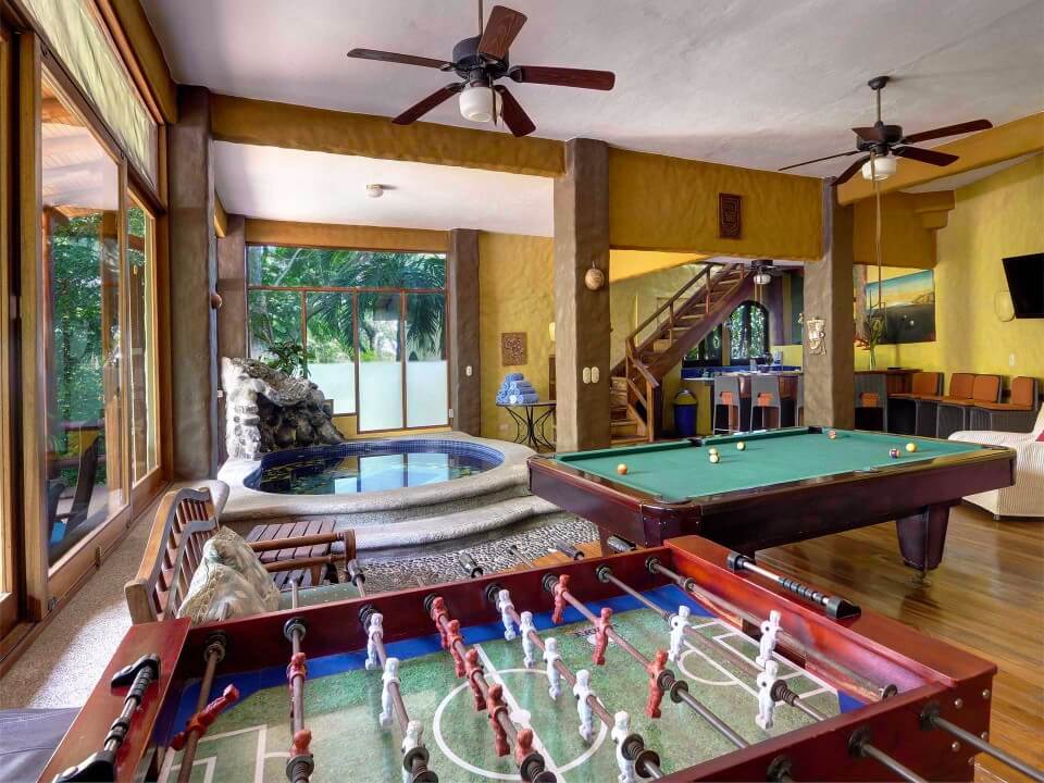 This large game lounge includes an indoor splash pool with a rock waterfall, a pool table, a foosball table, and a wet bar, with natural light from expansive windows framing jungle views.