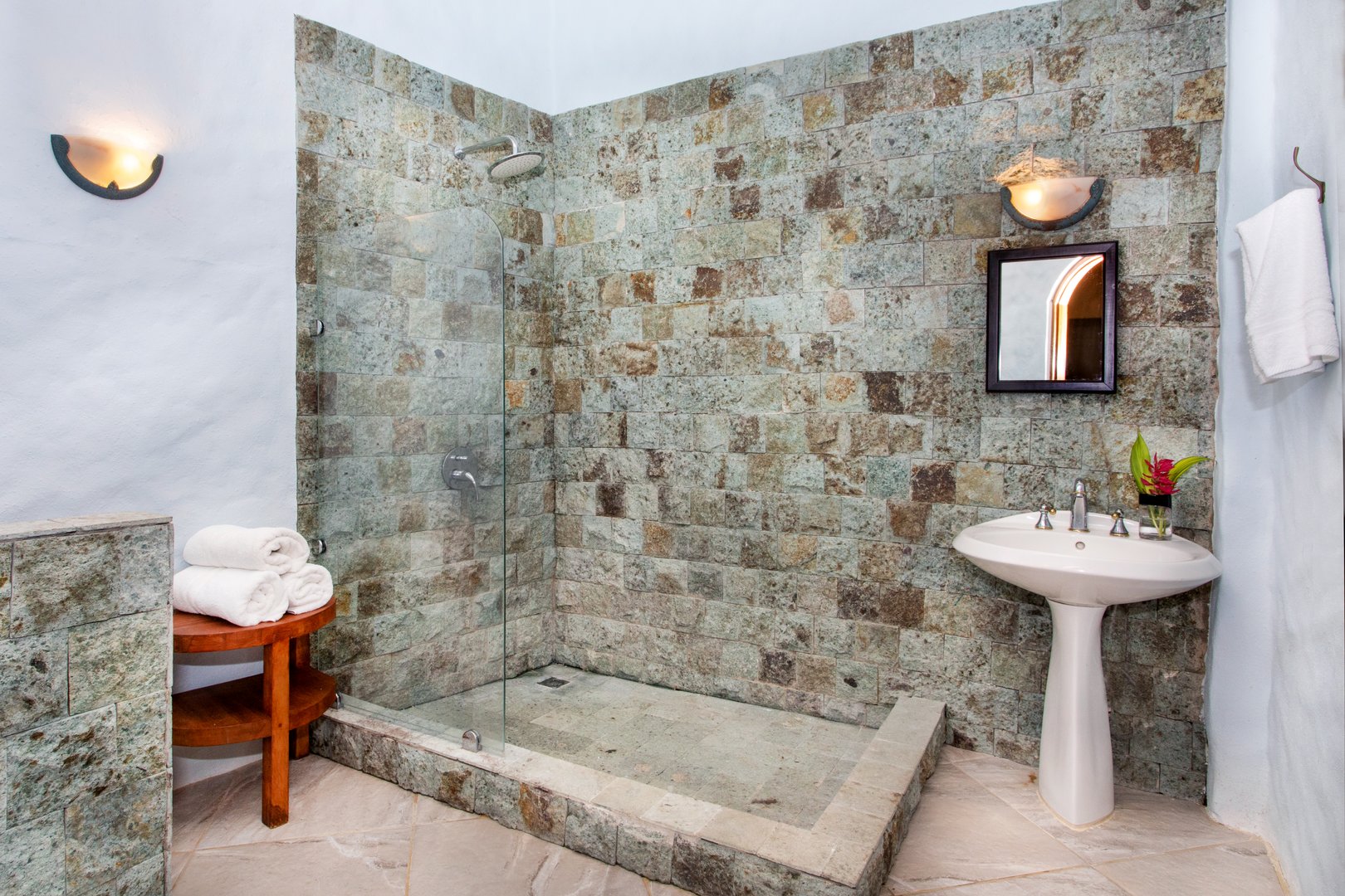 This bathroom features a walk-in shower with natural stone walls and floor, a white pedestal sink, and warm wall sconce lighting.