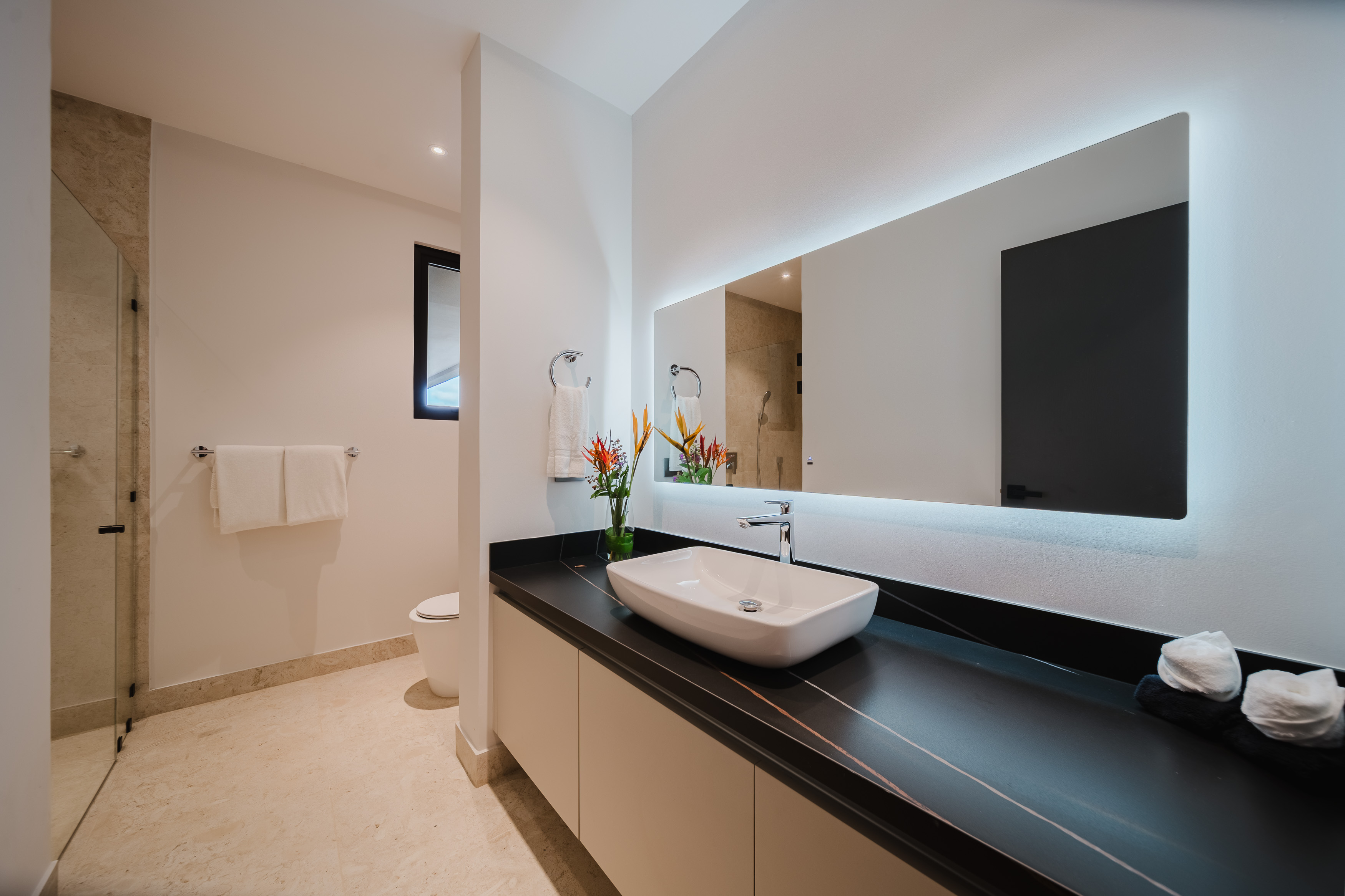 A bright, modern bathroom features a sleek vanity with a vessel sink, a large backlit mirror, and a walk-in shower with a glass enclosure.