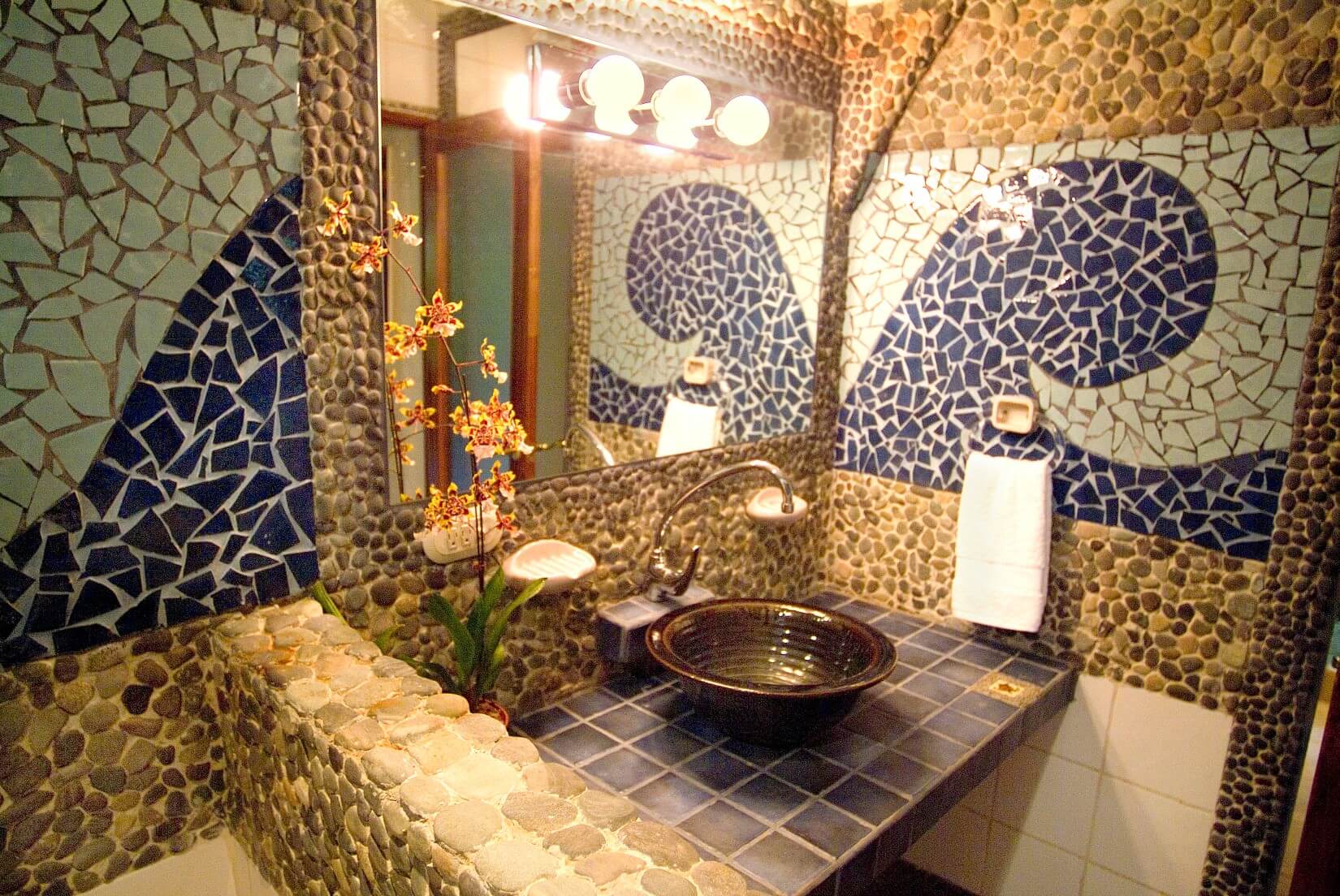 The bathroom features walls adorned with natural river stone mosaics and blue and white broken tile wave patterns, surrounding a dark vessel sink and mirror.