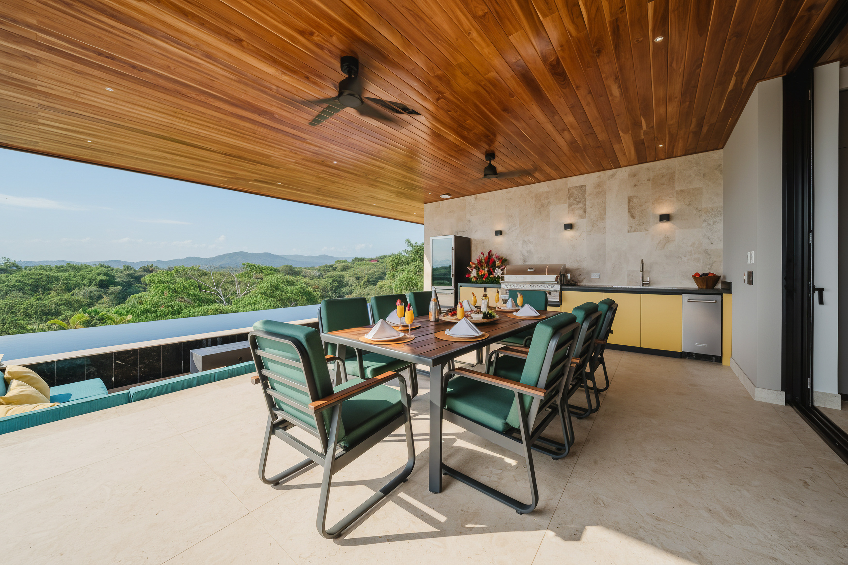 A spacious covered terrace with a wood-paneled ceiling features an outdoor dining area and kitchen, opening to an infinity pool and expansive views of the tropical landscape.