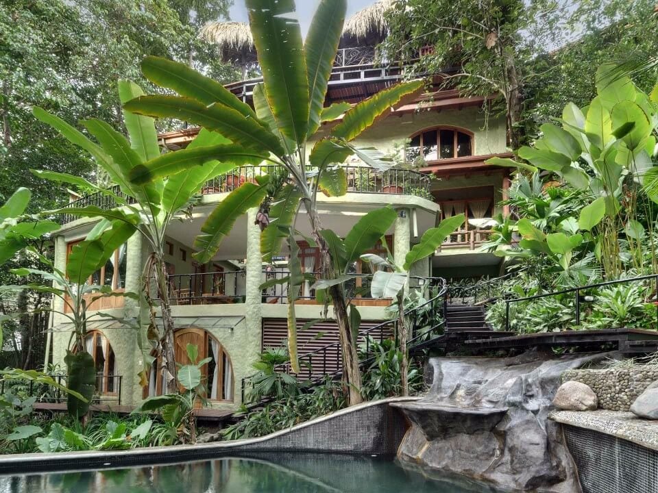 A multi-level villa, with light green walls and balconies, rises from dense tropical jungle, featuring a grotto-style pool with a rock waterfall in the foreground.
