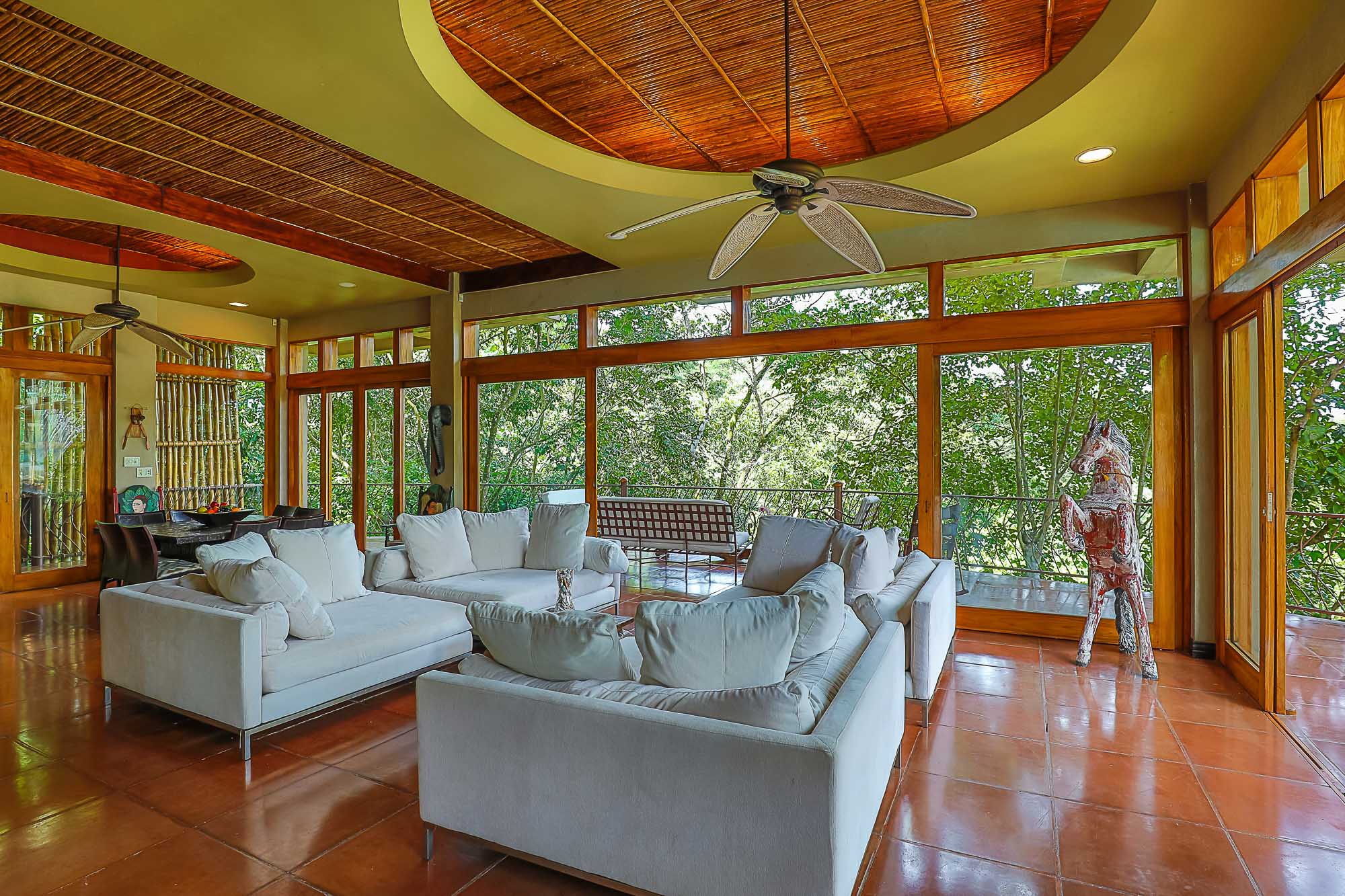 An open living area with comfortable white sofas and a bamboo ceiling is illuminated by natural light through expansive windows offering direct views of the surrounding rainforest.