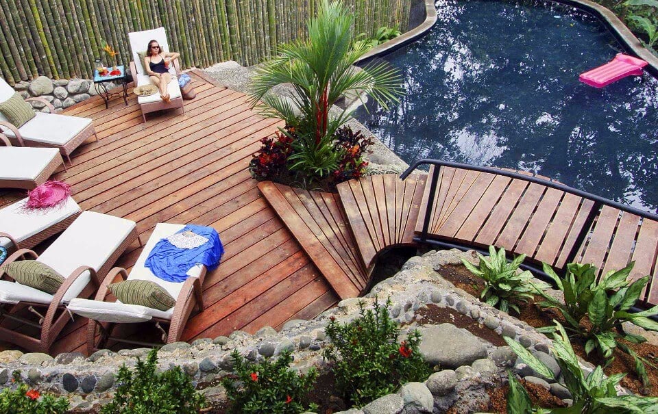A tropical outdoor pool area features a dark-bottomed pool, a large wooden sun deck with multiple lounge chairs, and lush natural landscaping providing privacy.