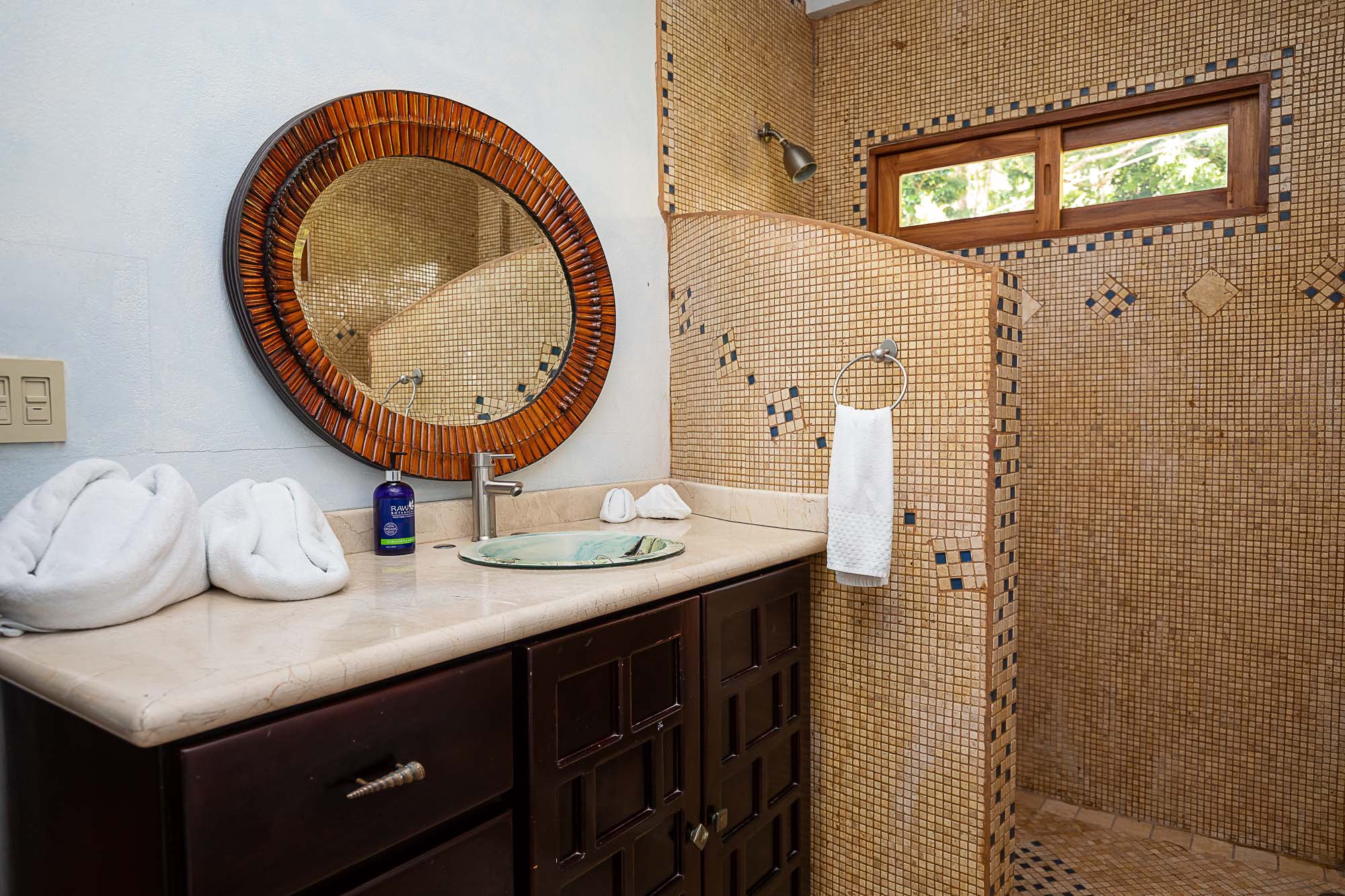 The bathroom features a dark wood vanity with a vessel sink and a large wood-framed mirror, complemented by a mosaic-tiled shower with a window that provides natural light and views of tropical greenery.