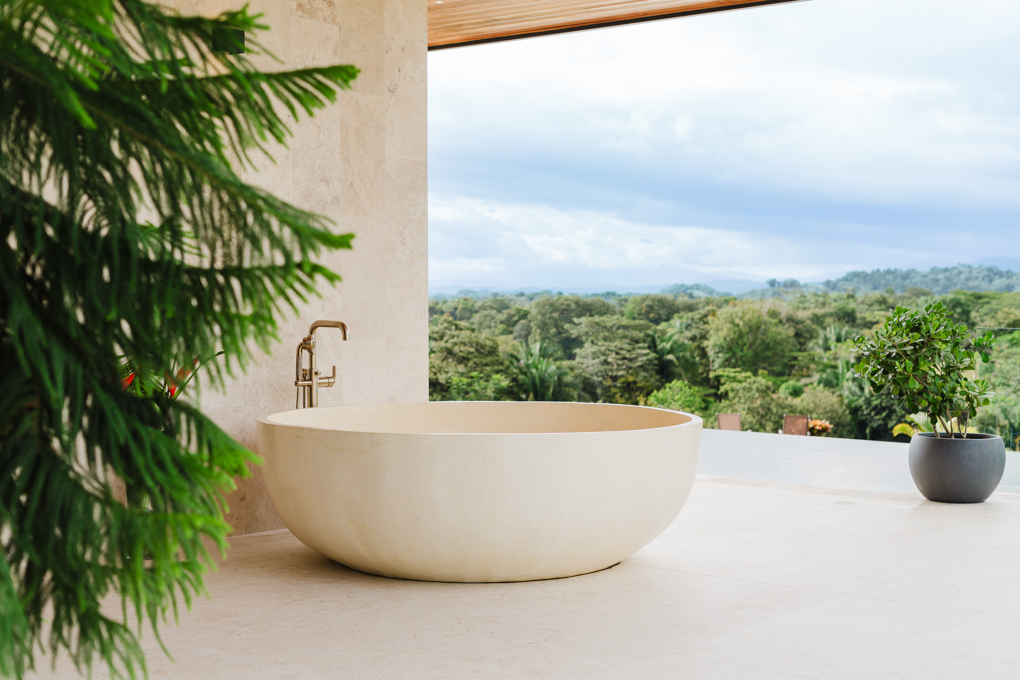 A large freestanding tub with a brass faucet is positioned next to an open wall looking out to a sprawling jungle and mountain view.