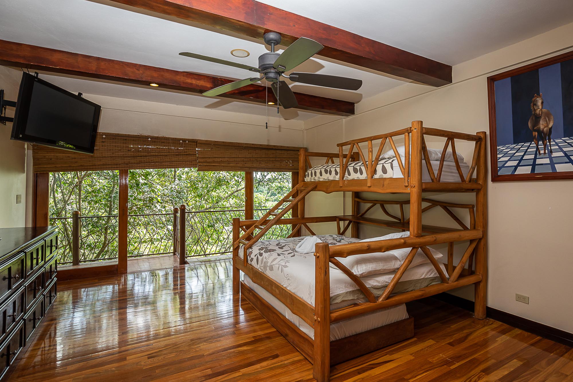 This bedroom features a sturdy wooden bunk bed, rich hardwood floors, and exposed ceiling beams, all bathed in natural light from the large window offering expansive rainforest views.