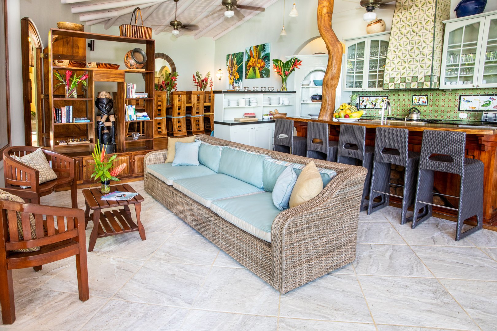 The open living area features a kitchen with a large wooden bar and counter seating, a lounge with a wicker sofa, and tropical hardwood furniture, all under a vaulted wood-beamed ceiling with a natural tree trunk column.