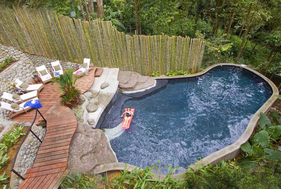 A freeform pool featuring a rock waterfall is surrounded by a curved wooden deck with lounge chairs and dense jungle foliage, creating a secluded outdoor space.