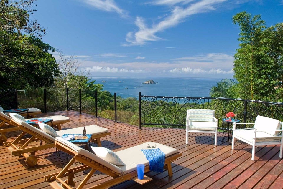 A sunlit wooden deck features multiple lounge chairs and armchairs, overlooking an expansive ocean vista framed by green jungle canopy.