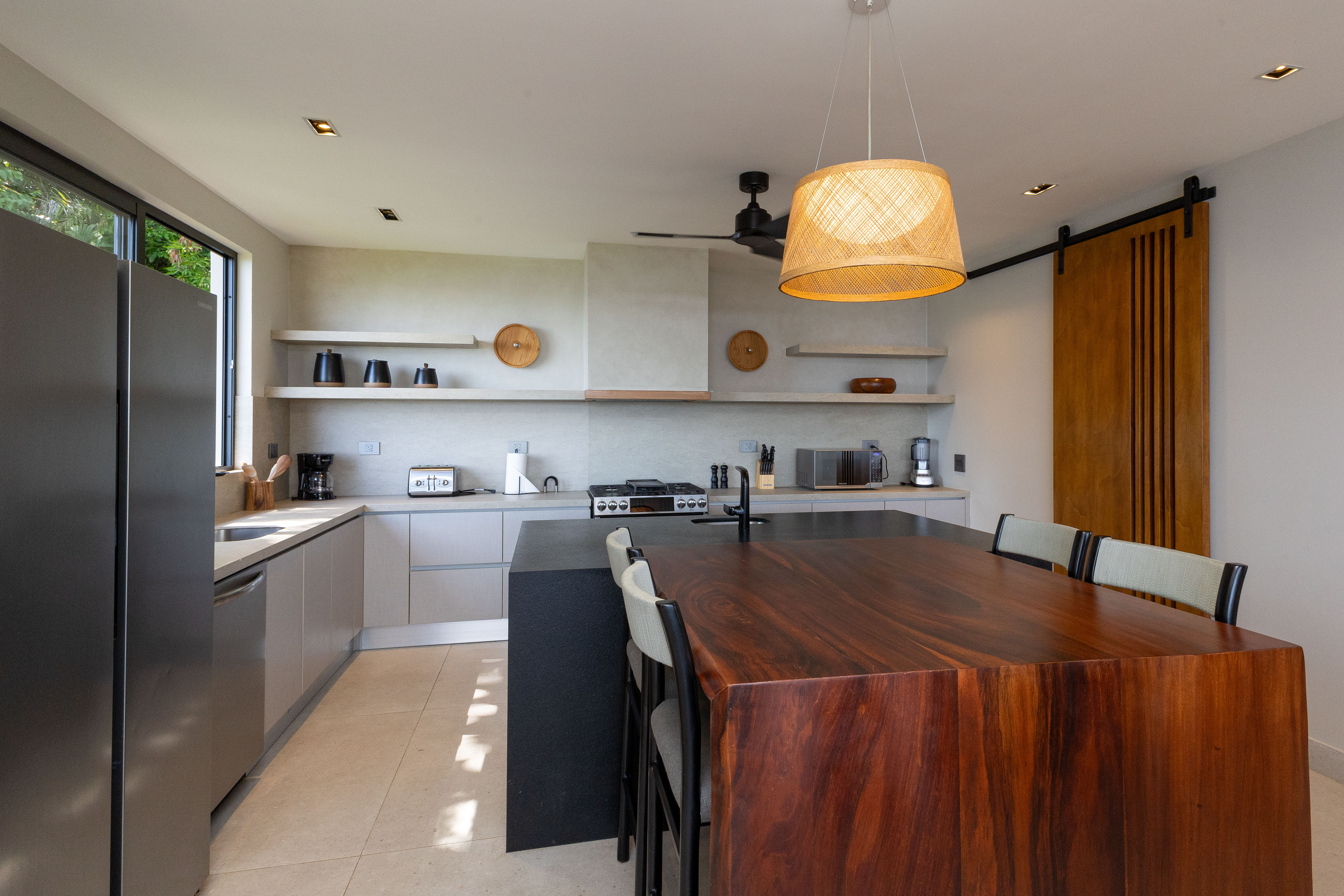This contemporary kitchen features an island with an integrated dining table, blending warm wood and dark stone, with a window offering natural light and jungle views.