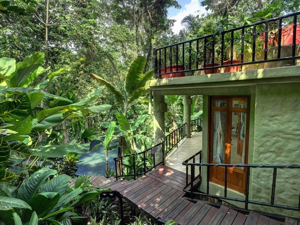 A wooden walkway descends through dense tropical foliage towards a sunlit jungle pool, next to a light green villa structure with a wooden door and an upper balcony.