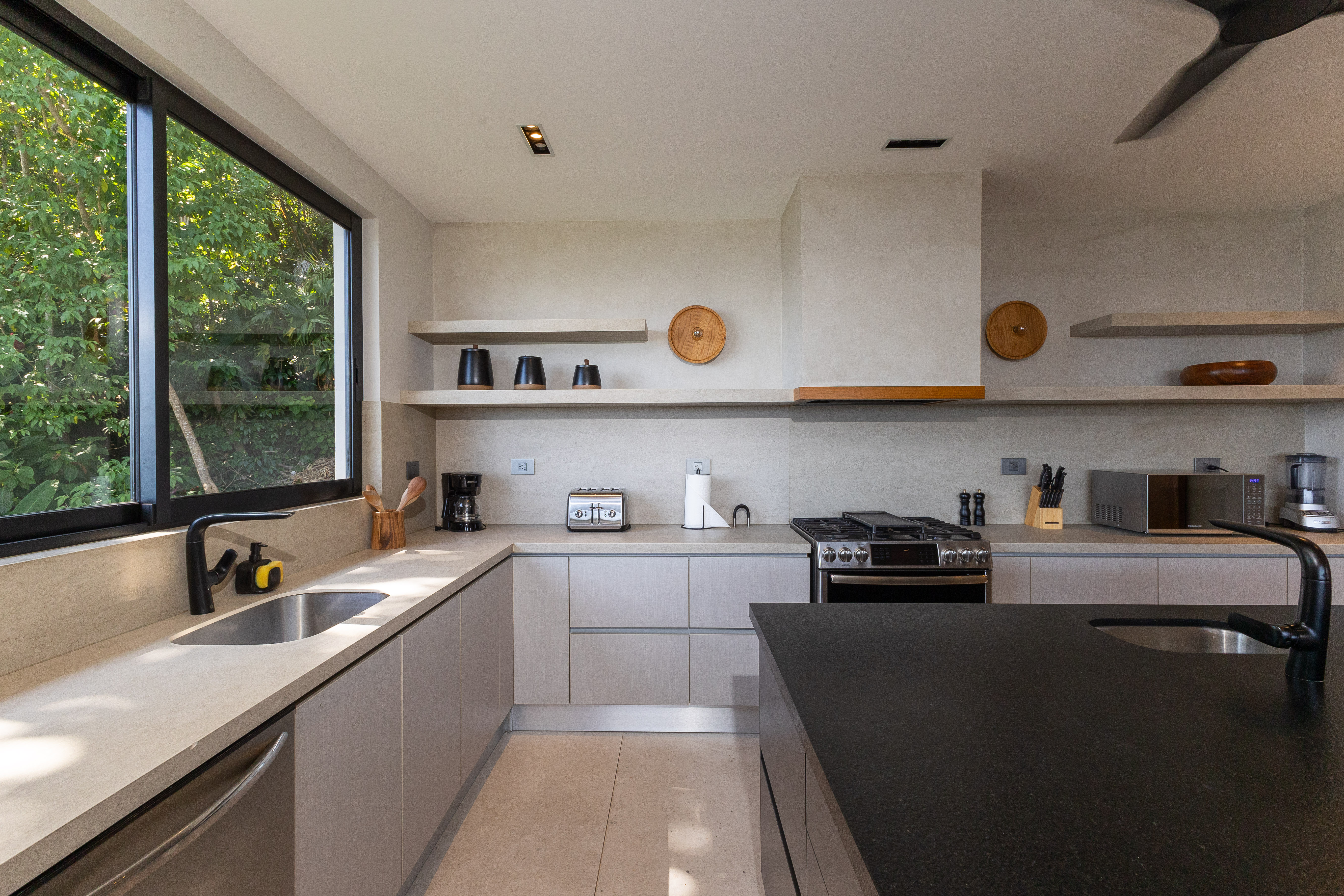A contemporary kitchen offers sleek light cabinetry, a dark island with a sink, modern appliances, and a large window overlooking lush jungle foliage.