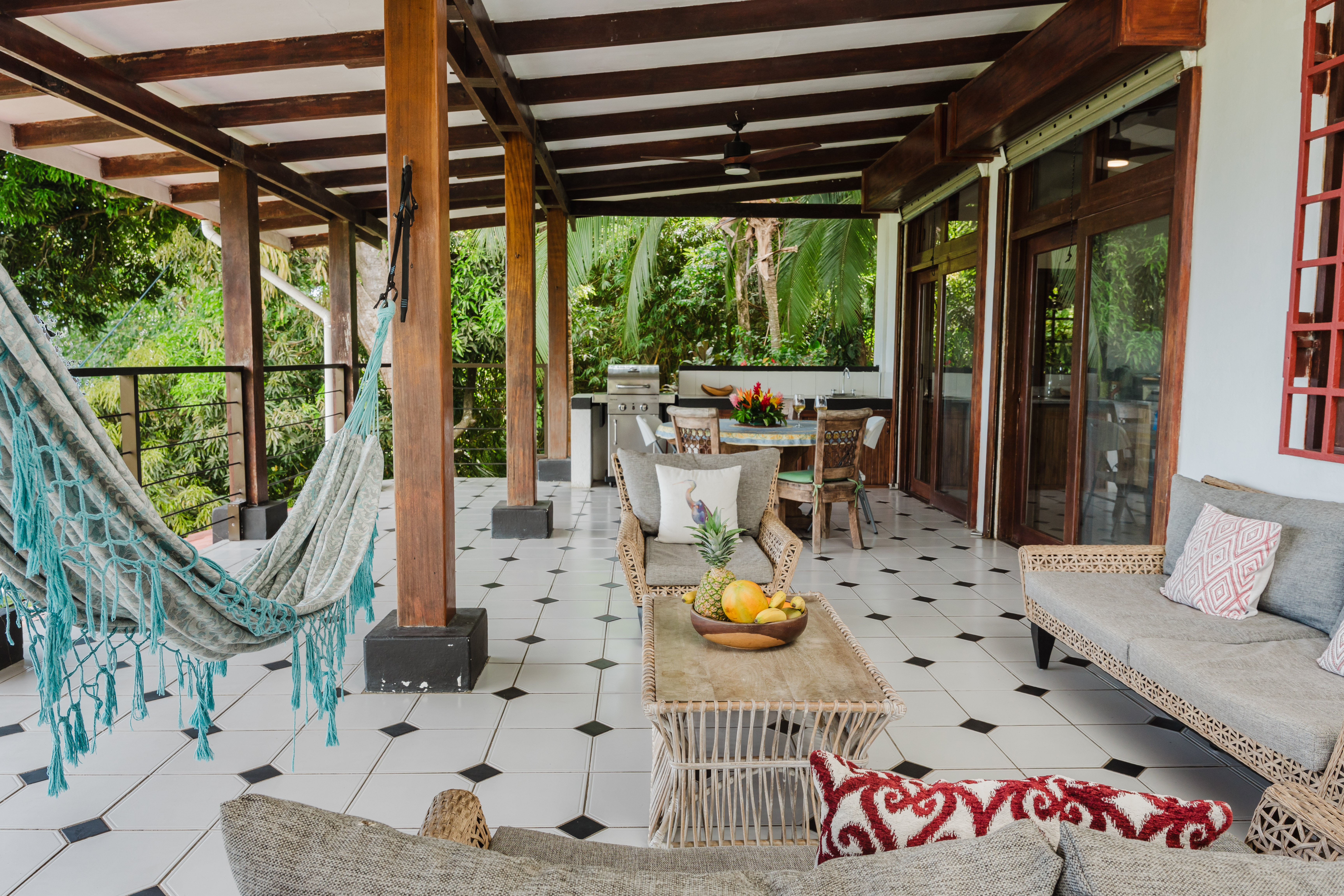 This spacious, covered outdoor terrace features an open-air living and dining area with wooden structural elements, tiled flooring, and views of tropical foliage.