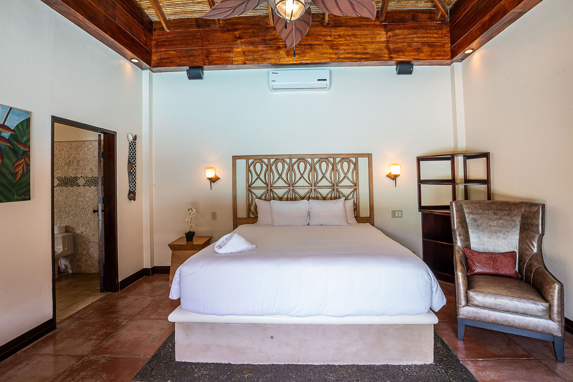 A spacious bedroom features a king bed with a carved wooden headboard, a high ceiling with exposed wooden beams and a natural woven finish, and an adjacent en-suite bathroom.