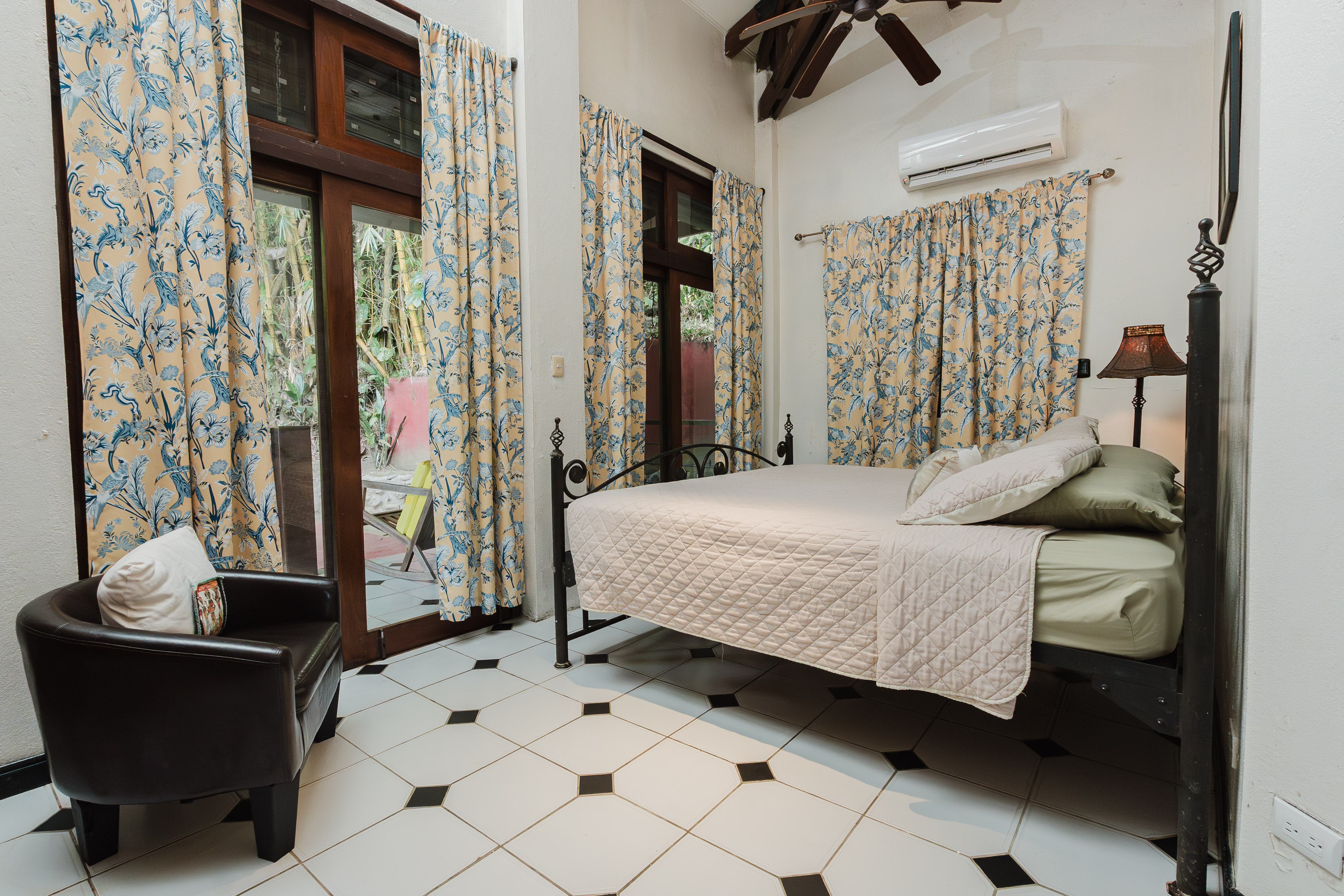 An air-conditioned bedroom features a wrought iron bed, black and white tiled floor, exposed wooden ceiling beams, and large glass doors providing natural light and a view of tropical foliage.