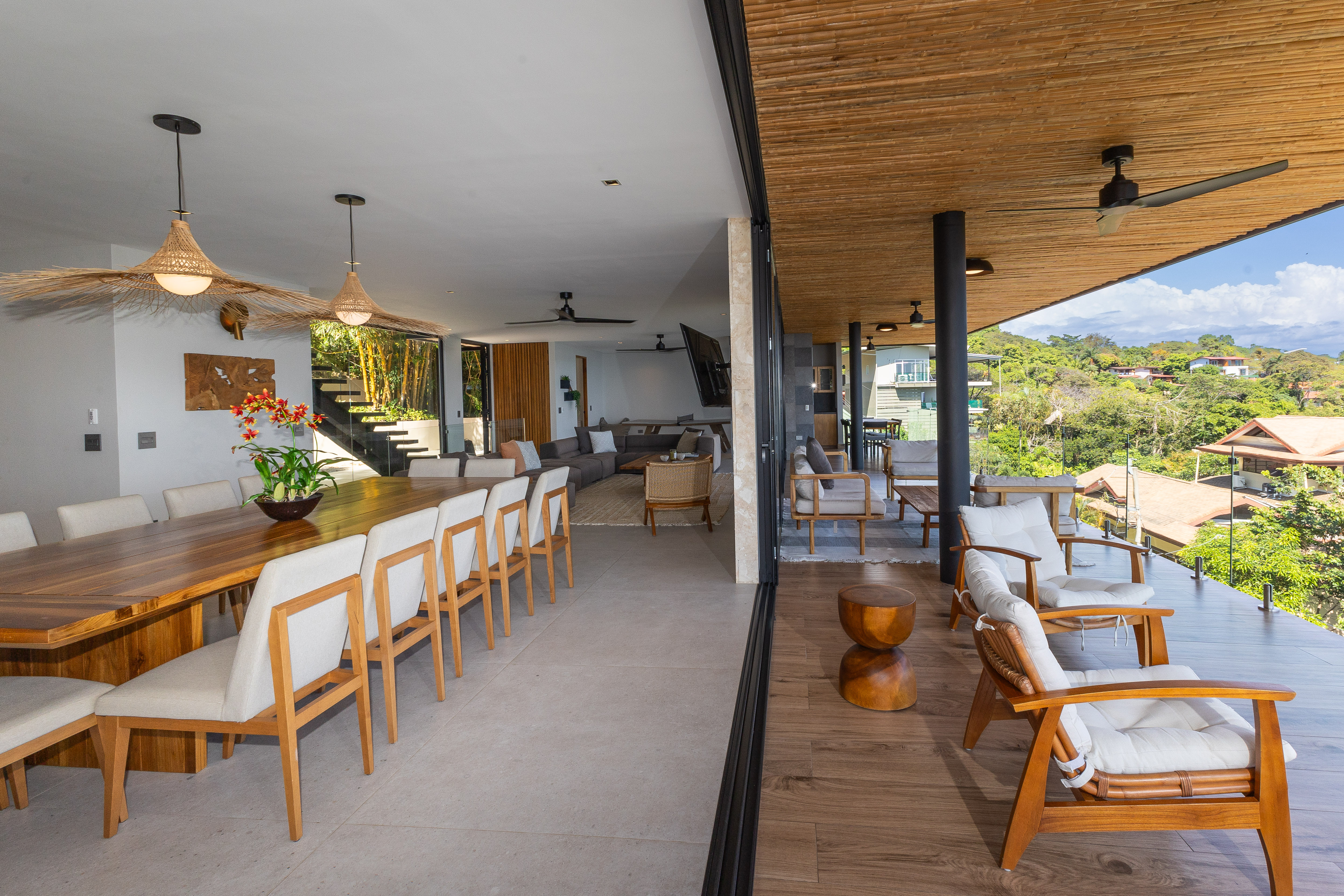 A spacious outdoor terrace with natural wood lounge furniture under a bamboo ceiling provides open views of tropical hills and blends seamlessly with an adjacent indoor dining and living area.