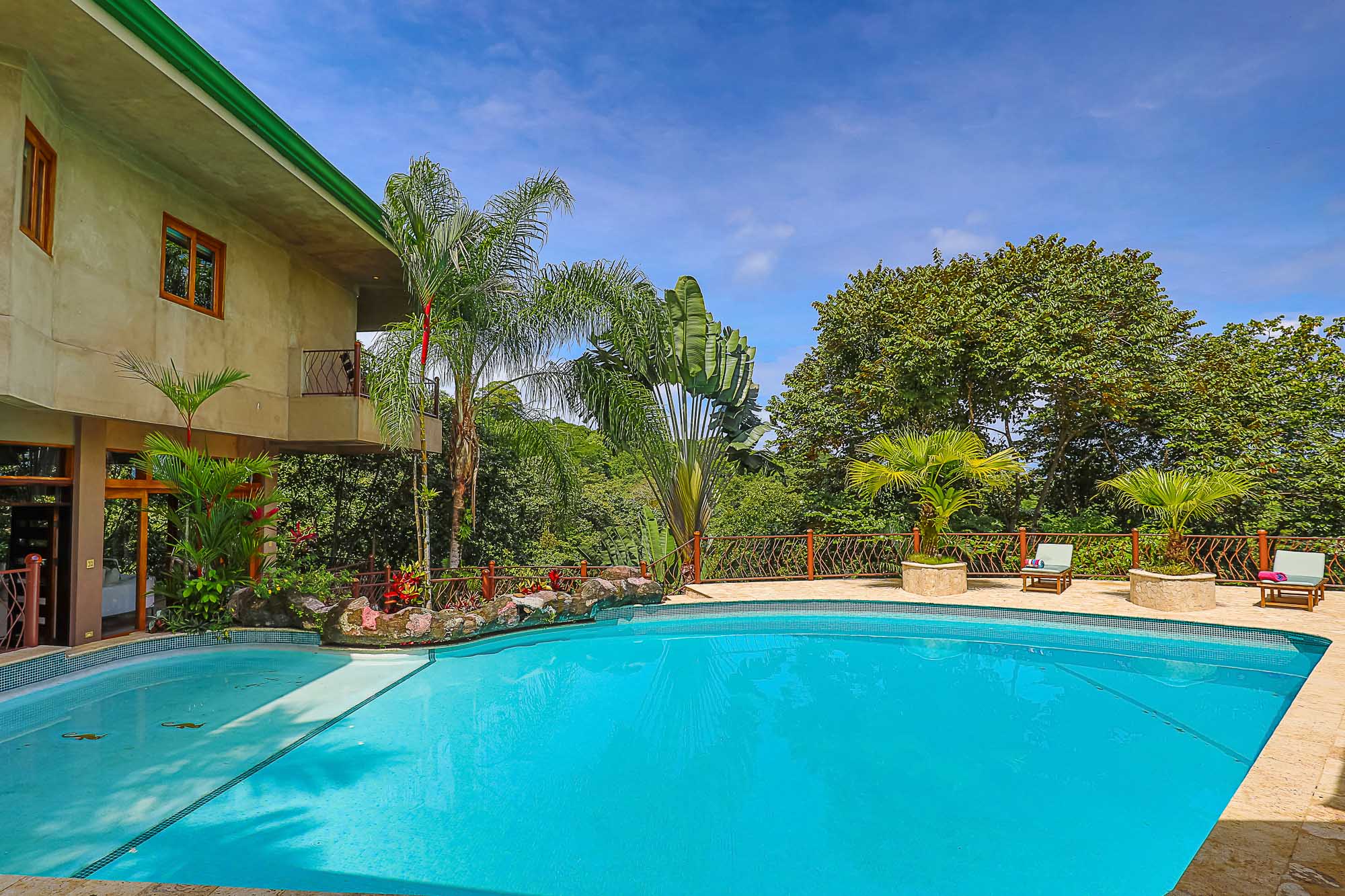 A large blue swimming pool is set against a multi-story villa, surrounded by tropical forest greenery under a bright blue sky.