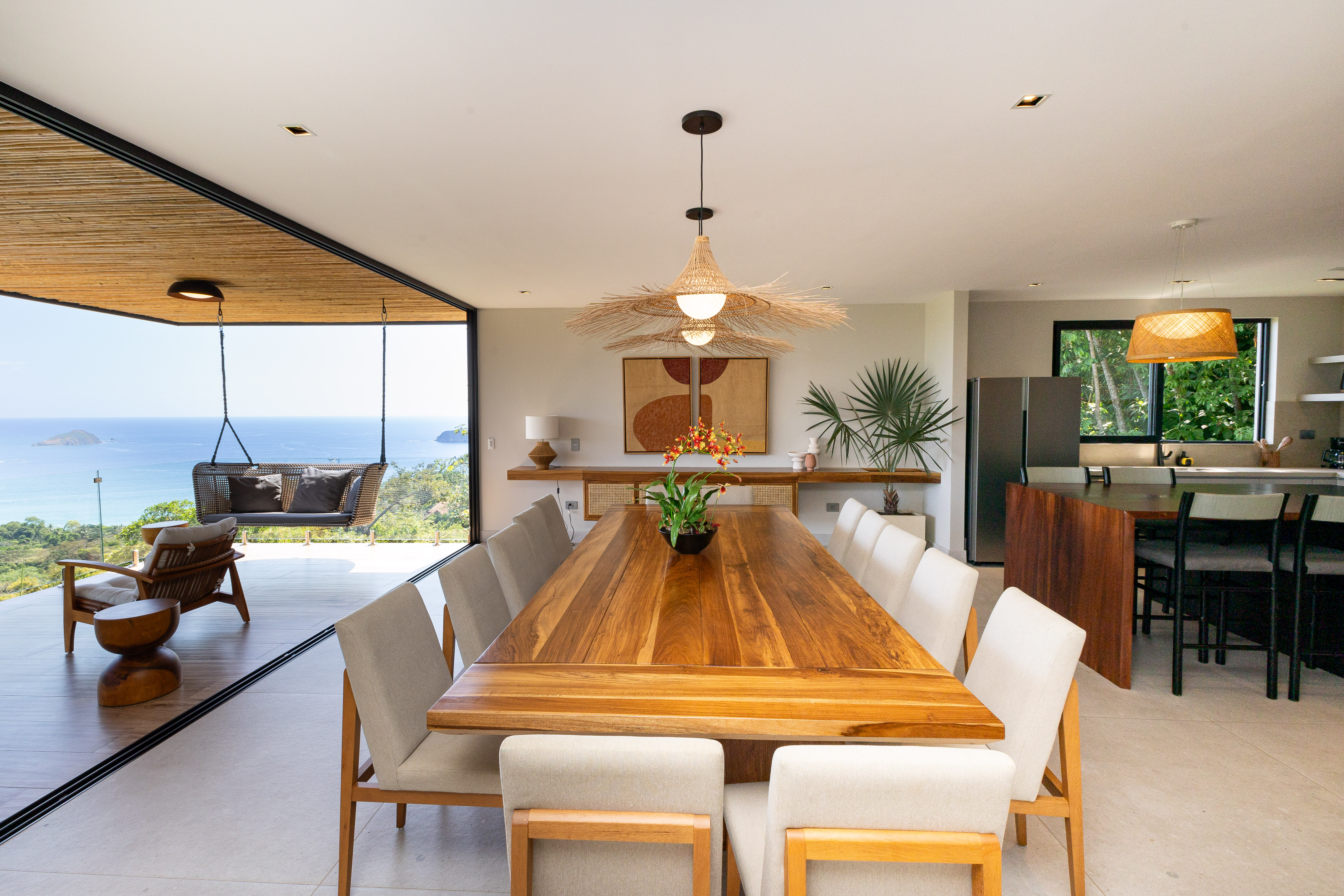 An open-plan dining space, featuring a long wooden table and numerous chairs, is bathed in natural light from full-height glass doors that lead to a terrace with broad ocean and island views.