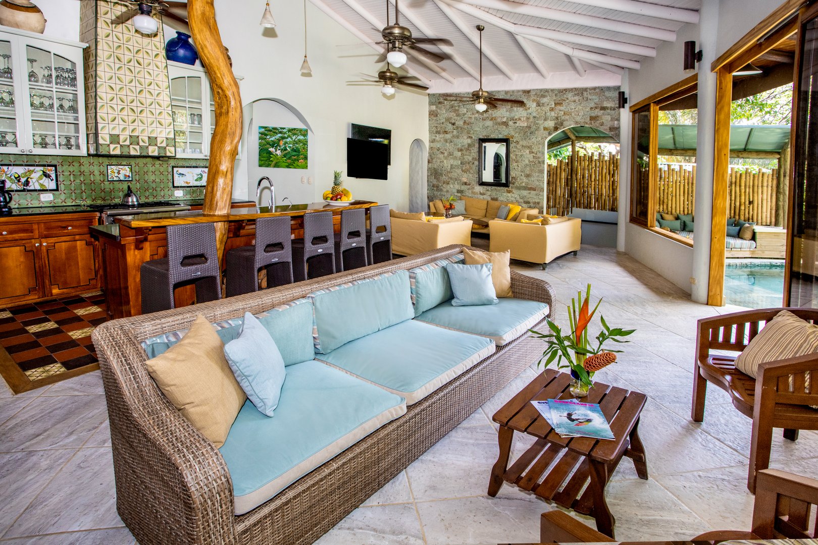 This open-plan living area combines a kitchen with bar seating and multiple lounge zones under a high vaulted ceiling, opening to a pool area.