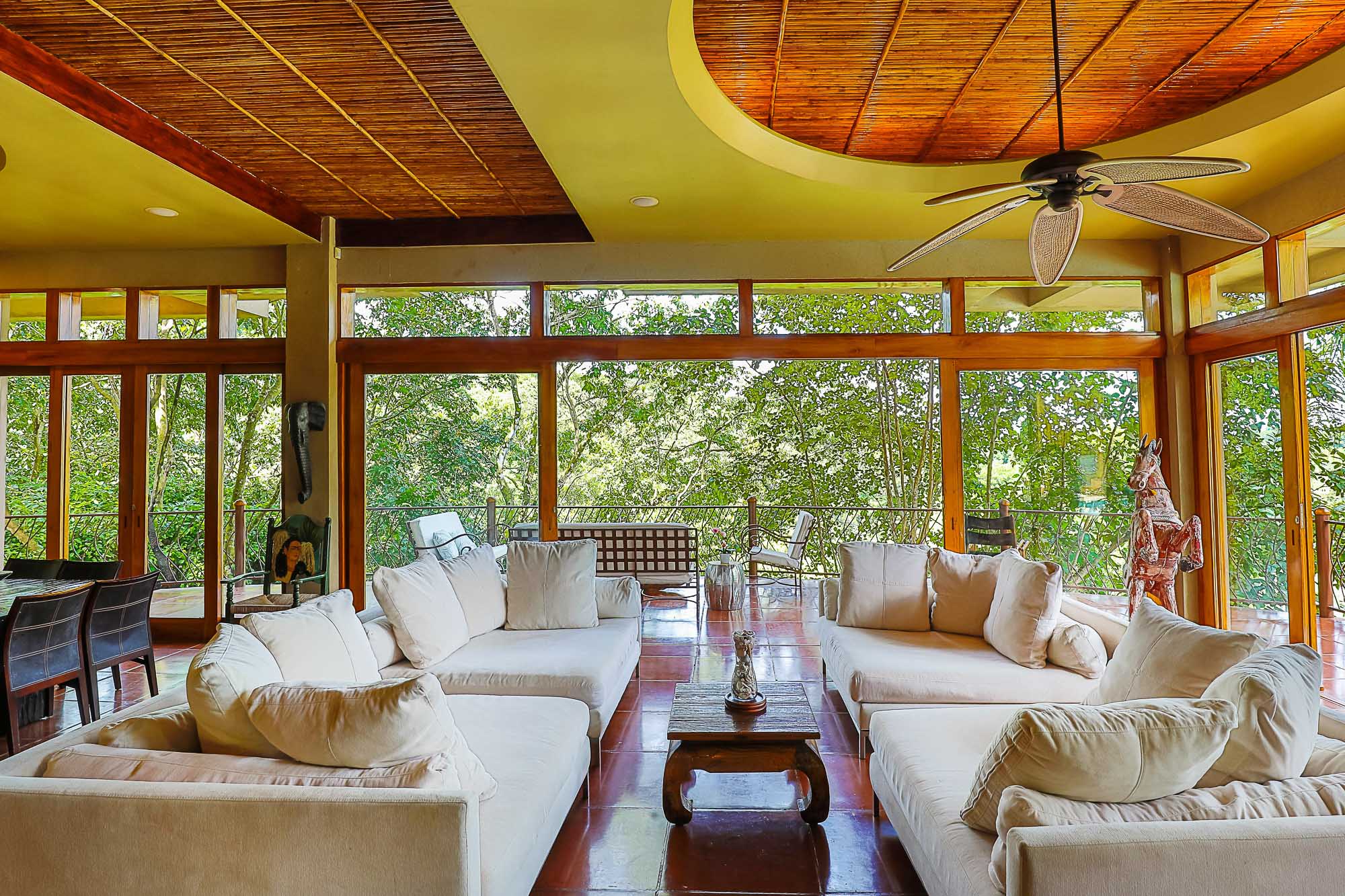 The living room features multiple large sofas, a natural wood paneled ceiling, and a wall of windows offering direct views of the tropical rainforest and plentiful natural light.