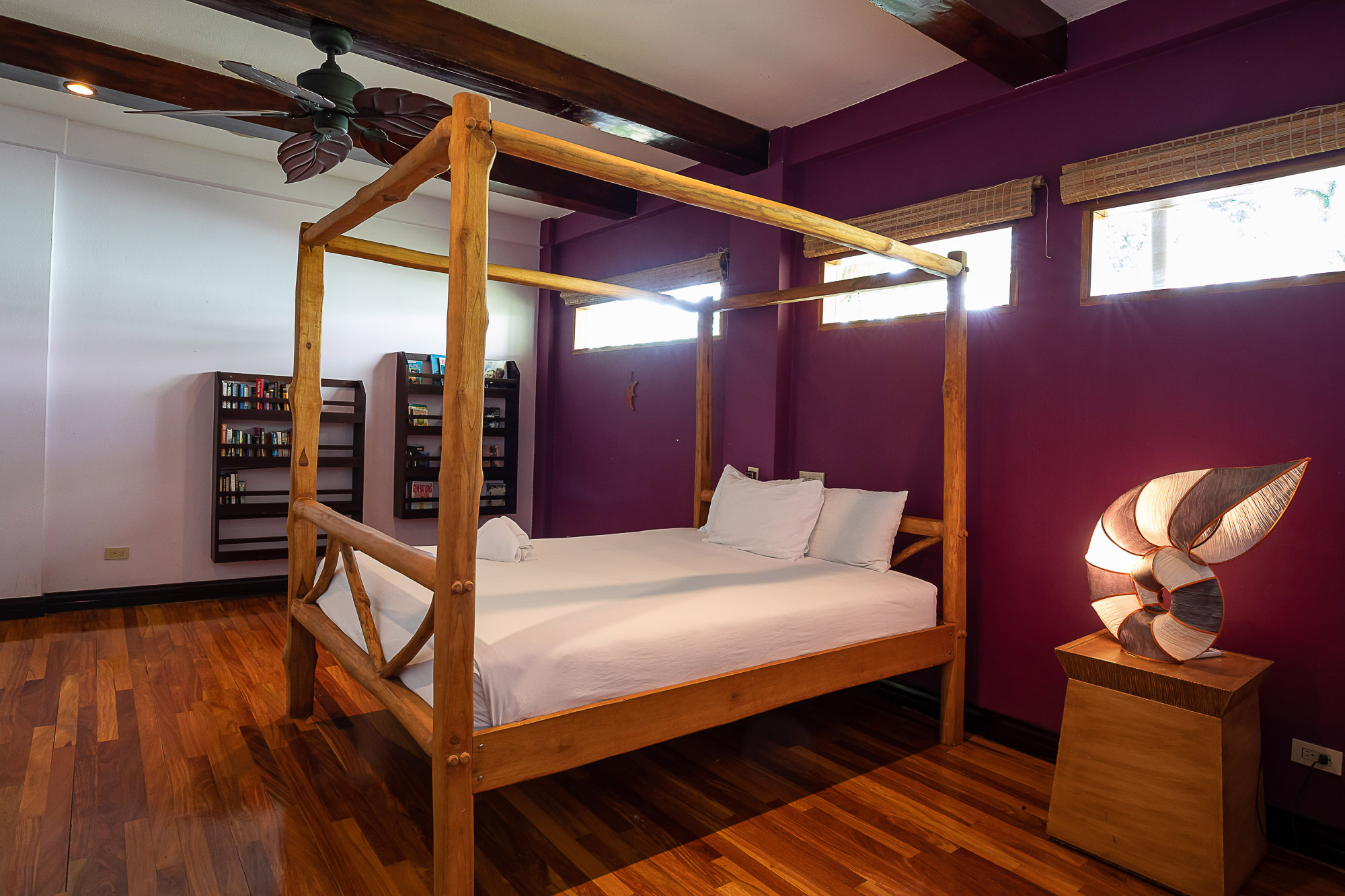 This bedroom features a rustic four-poster wooden bed, polished hardwood floors, dark ceiling beams, and natural light entering through horizontal windows with woven blinds, creating a tropical retreat.