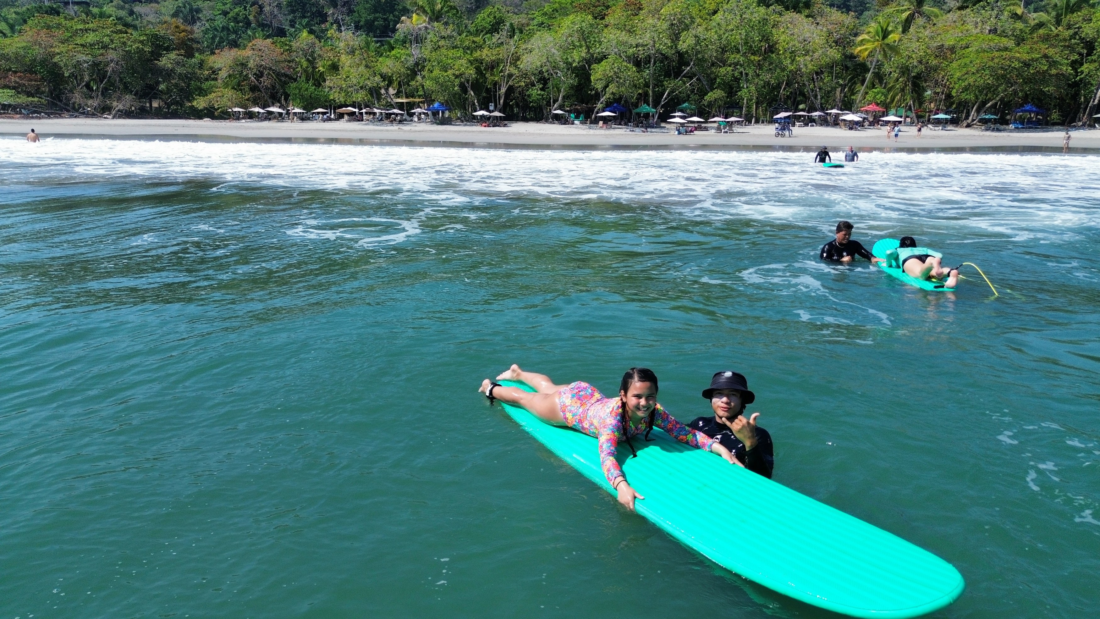 A sunny beach scene shows people in clear ocean water learning to surf, with a sandy shore lined by numerous umbrellas and tropical trees.