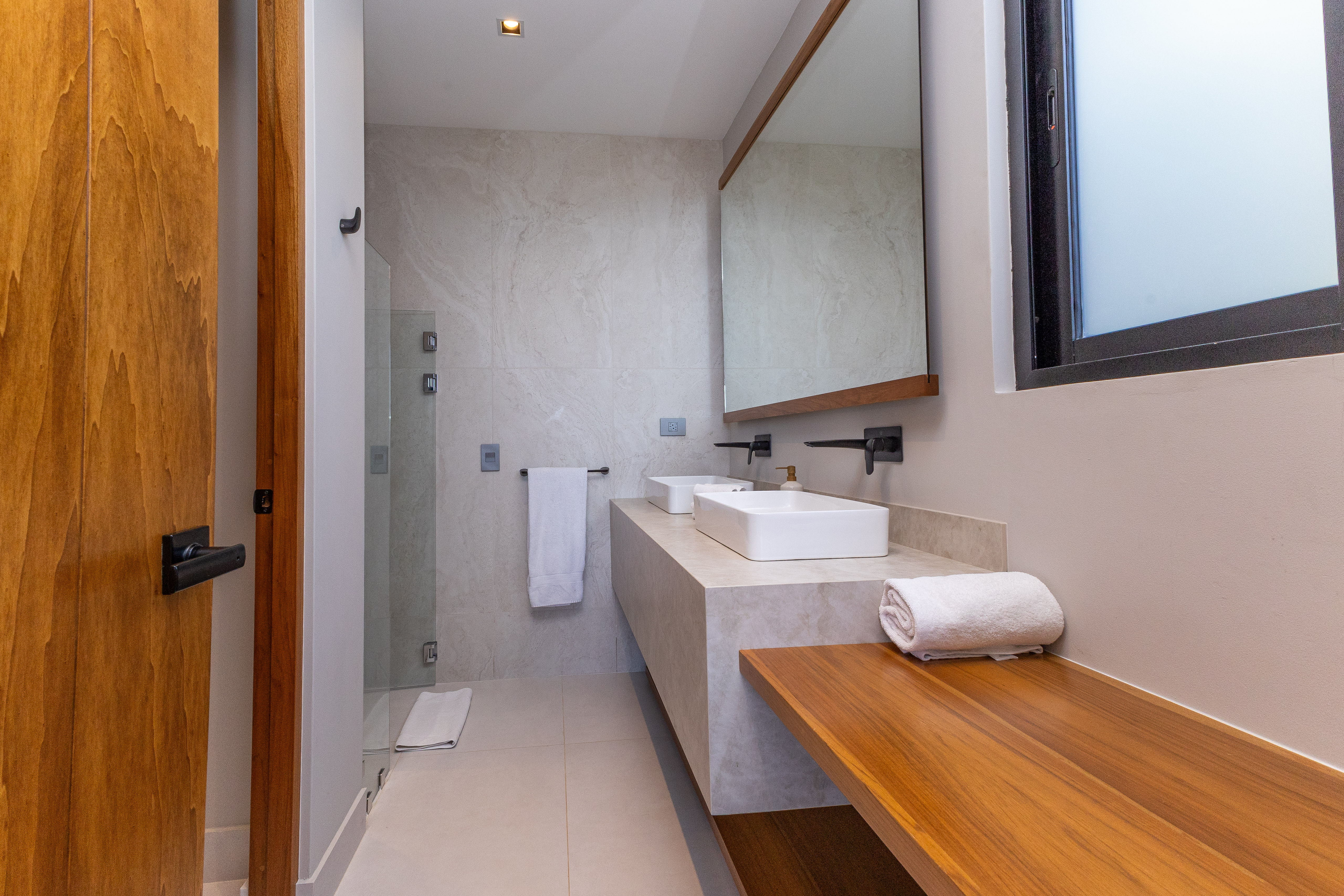 This bright bathroom features a modern double vanity with wall-mounted fixtures, a glass-enclosed shower, and warm wood details, designed with natural light from a window.