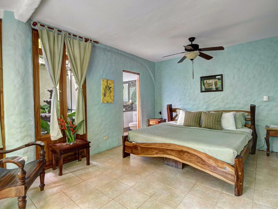 A light-filled bedroom features a natural wood bed frame, textured walls, tile flooring, and large glass doors opening to a garden fountain view, with an open doorway leading to a private bathroom.