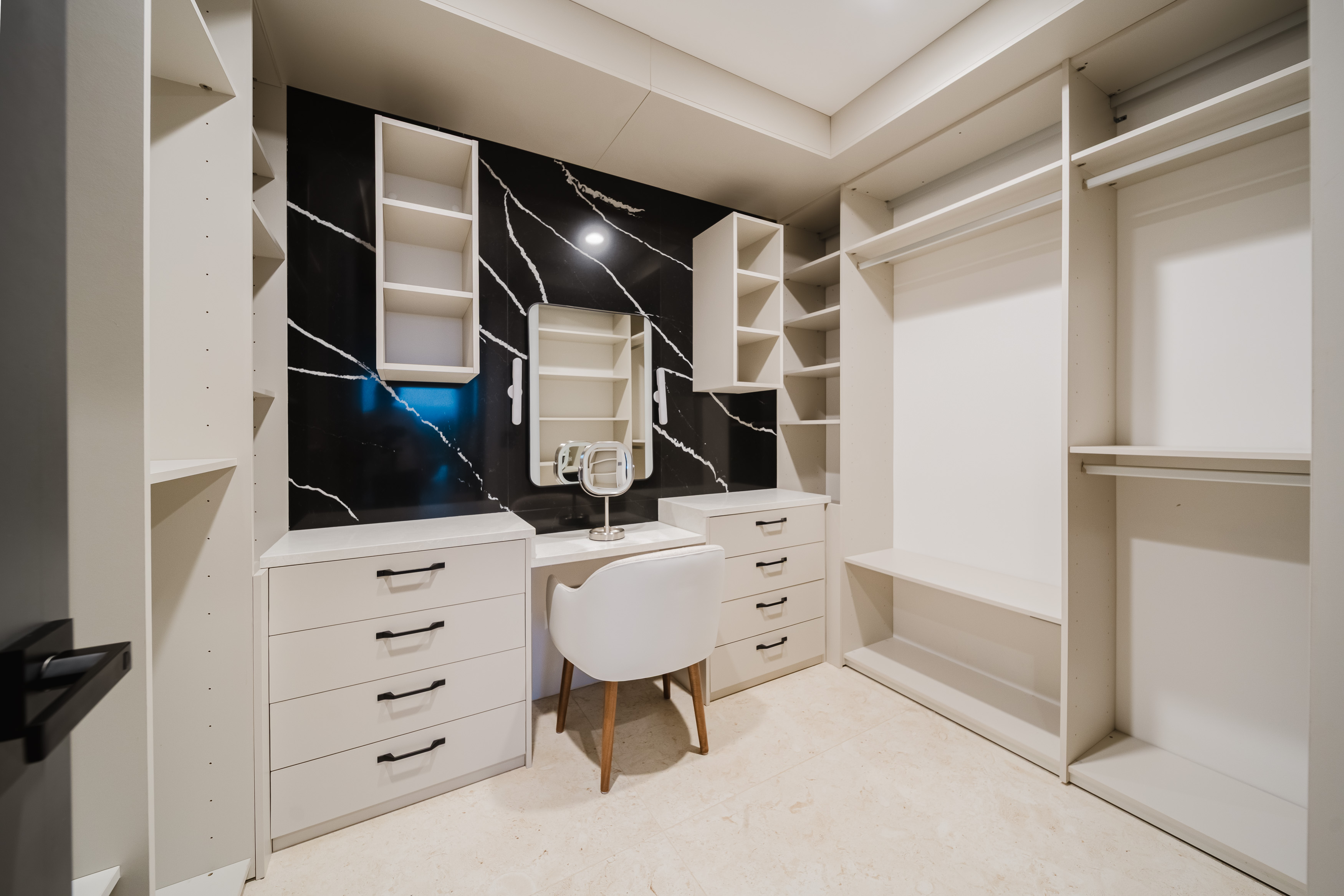 A dedicated dressing room features extensive built-in light-colored cabinetry and shelving, with a central vanity area set against a striking dark marble-patterned accent wall, illuminated by overhead lighting.