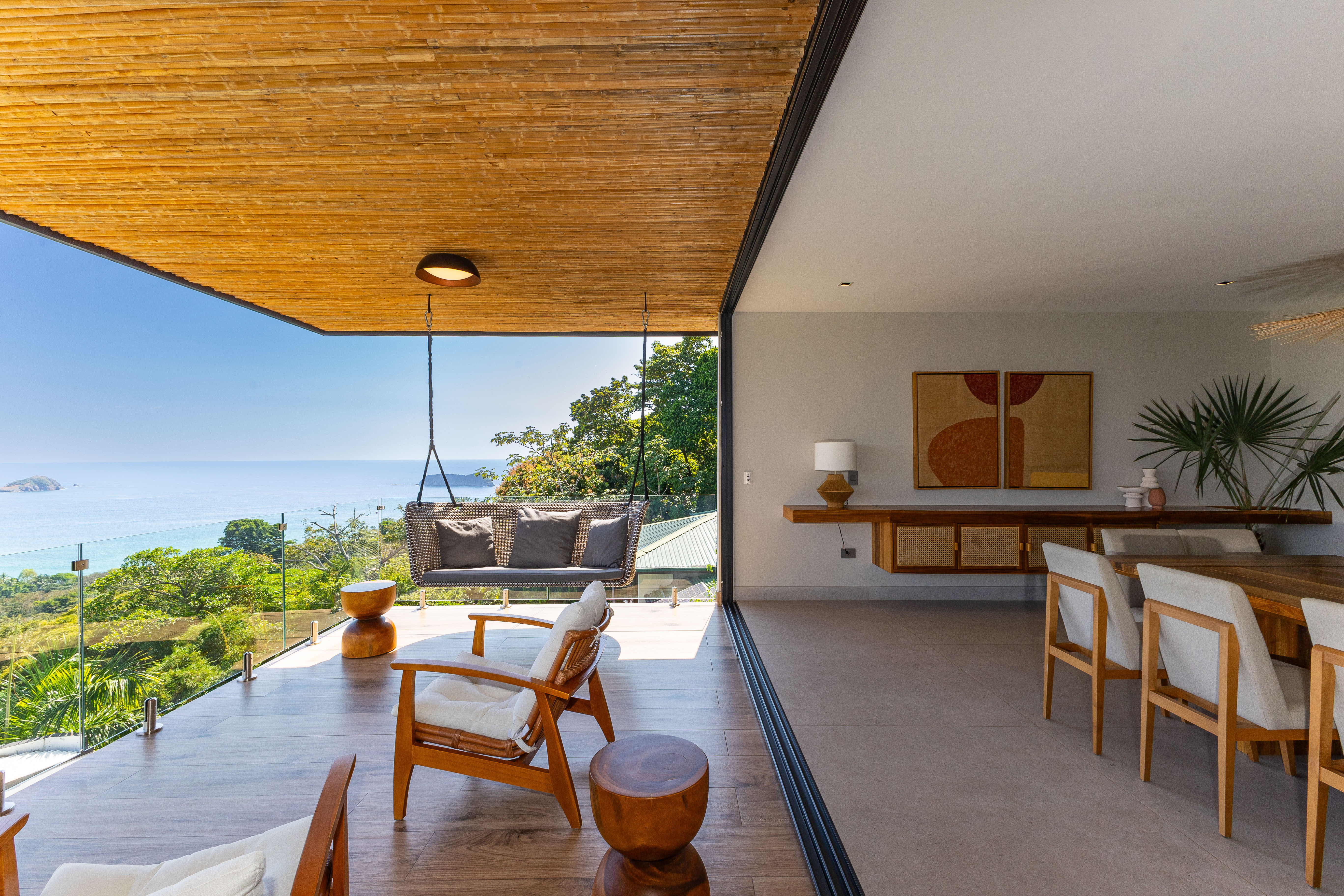 This open terrace, featuring a bamboo ceiling and multiple seating options including a hanging swing, provides direct access to indoor dining and frames expansive ocean and jungle views.