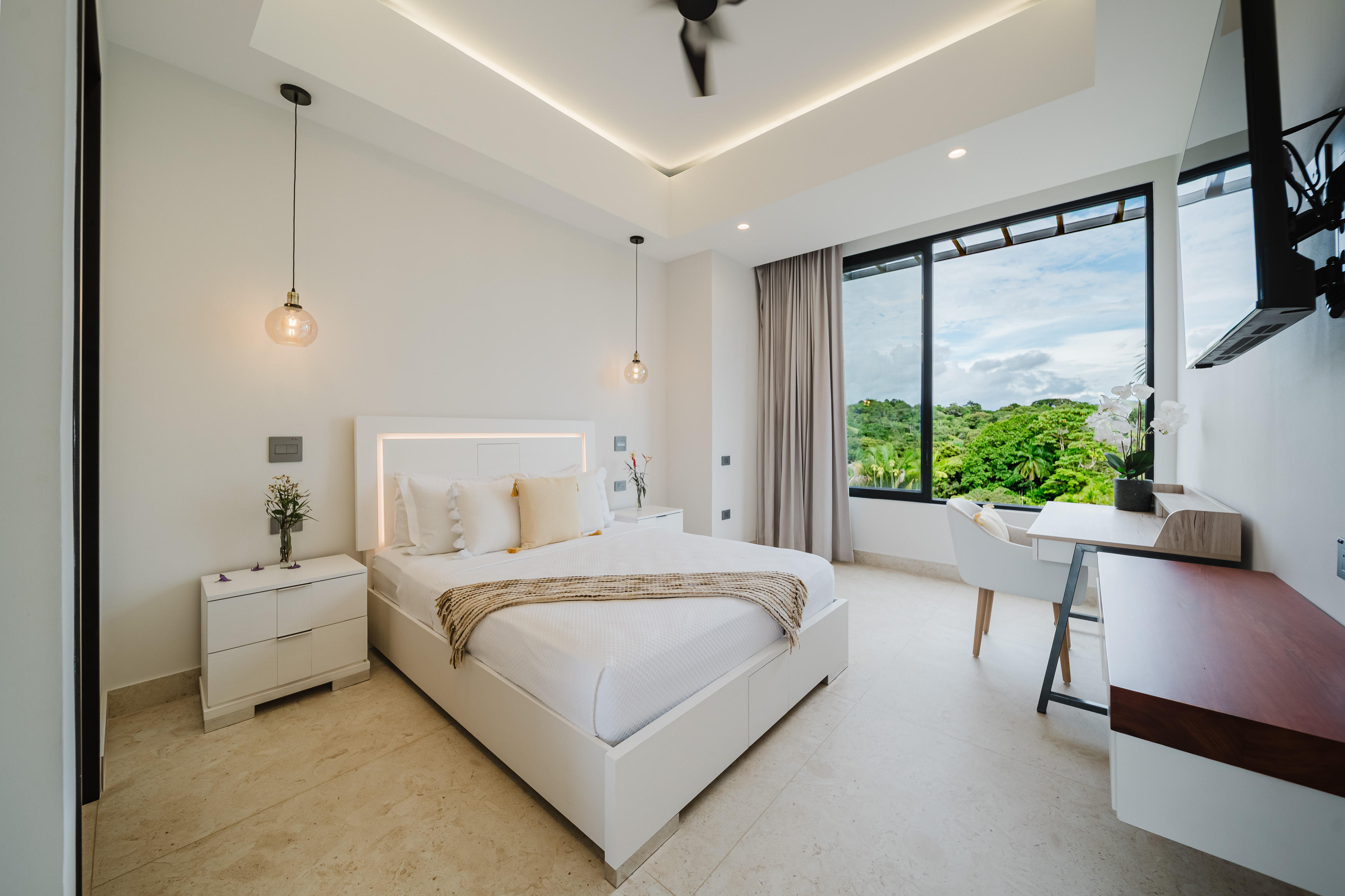 This bright bedroom features a modern white bed with a backlit headboard, illuminated by natural light from a large window offering expansive views of the surrounding tropical jungle.