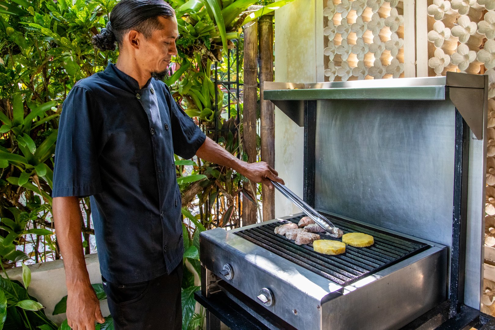 An outdoor stainless steel grill is positioned among lush tropical foliage, complemented by decorative wall panels and natural light.