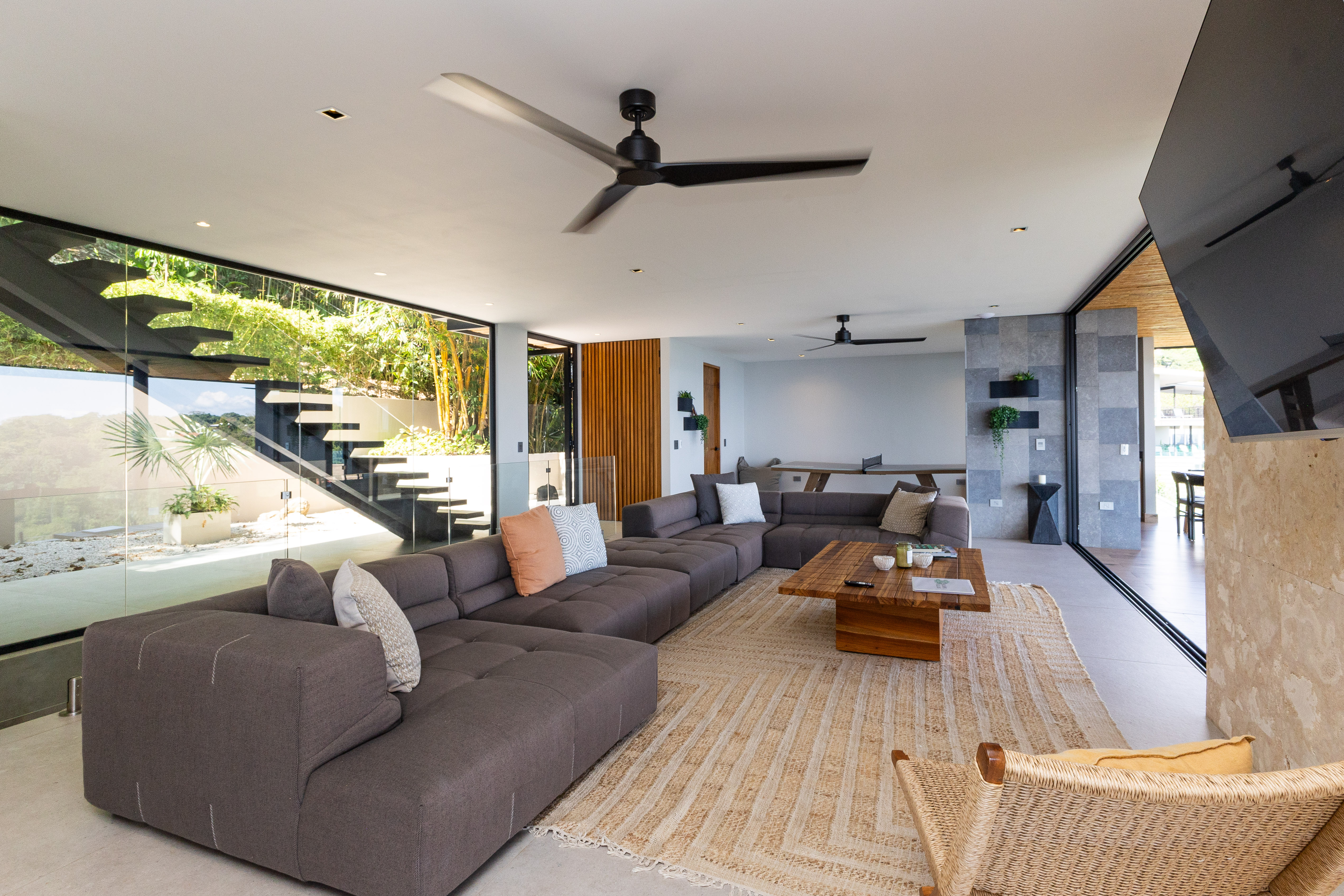 An expansive, light-filled living area features a large sectional sofa, floor-to-ceiling glass offering jungle views, and a nearby ping pong lounge.