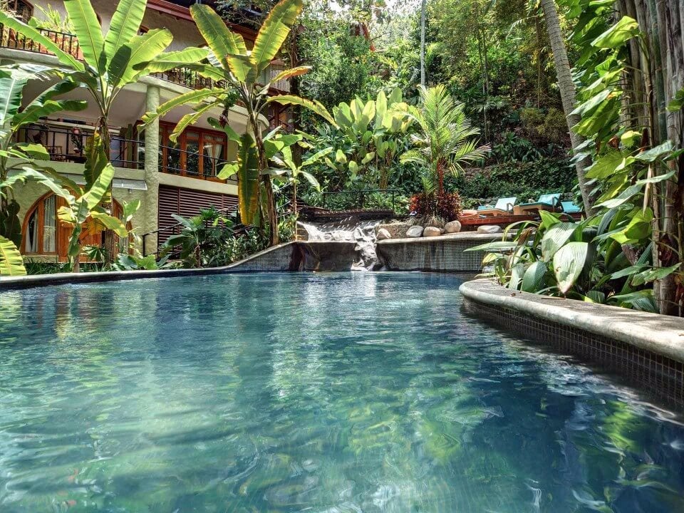 The tranquil jungle pool with a cascading waterfall sits before the villa's multi-level facade, enveloped by lush tropical foliage.
