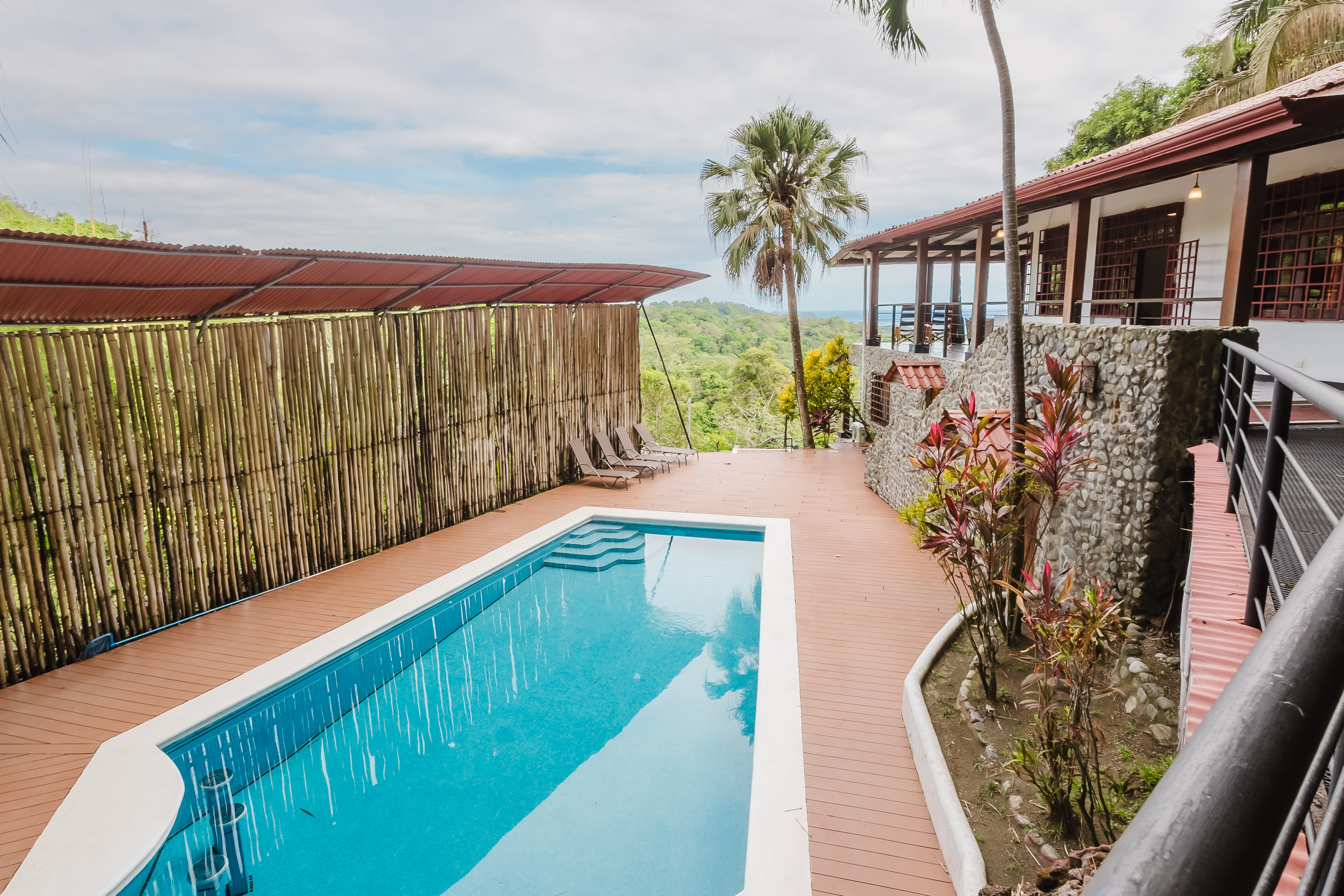 A private swimming pool with steps is surrounded by a wood deck and tropical landscaping, featuring a villa with a stone facade and an upper-level terrace overlooking distant ocean and lush jungle.