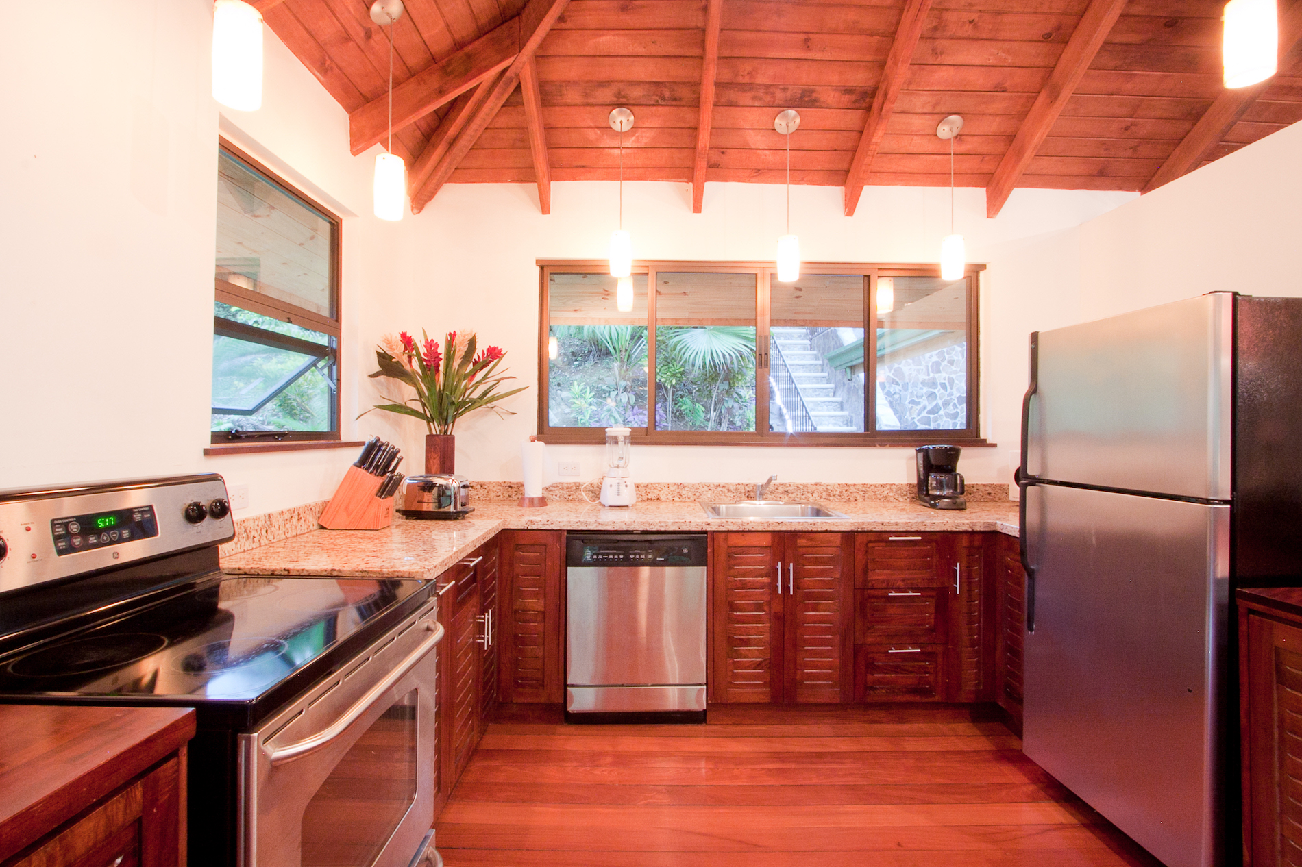 A well-appointed kitchen features rich wooden floors, cabinetry, and a vaulted ceiling, complemented by stainless steel appliances and windows overlooking the tropical jungle.