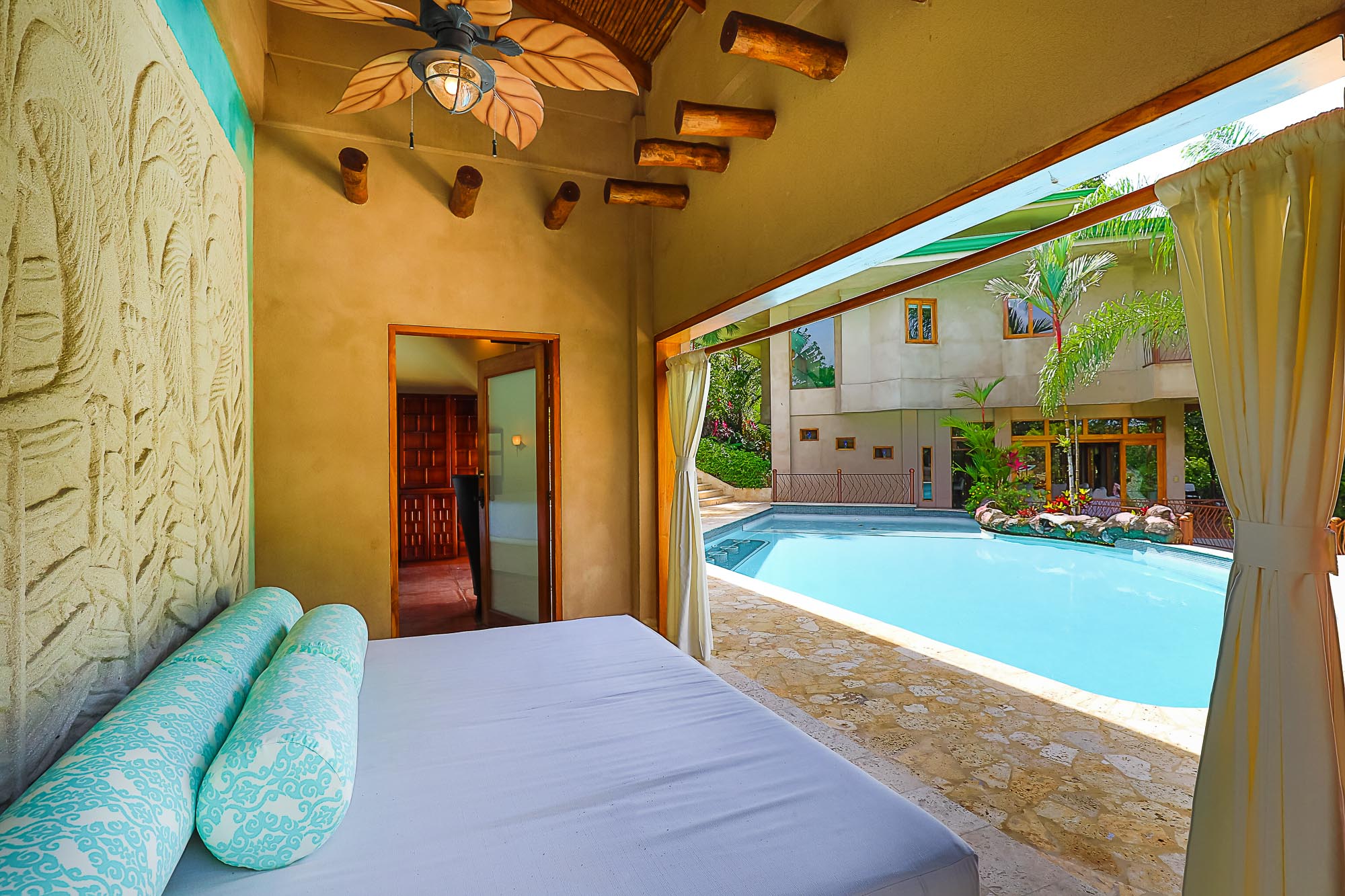 This covered outdoor lounge features a daybed, exposed wooden beams, and a leaf-bladed ceiling fan, with open views to the pool and the main villa.