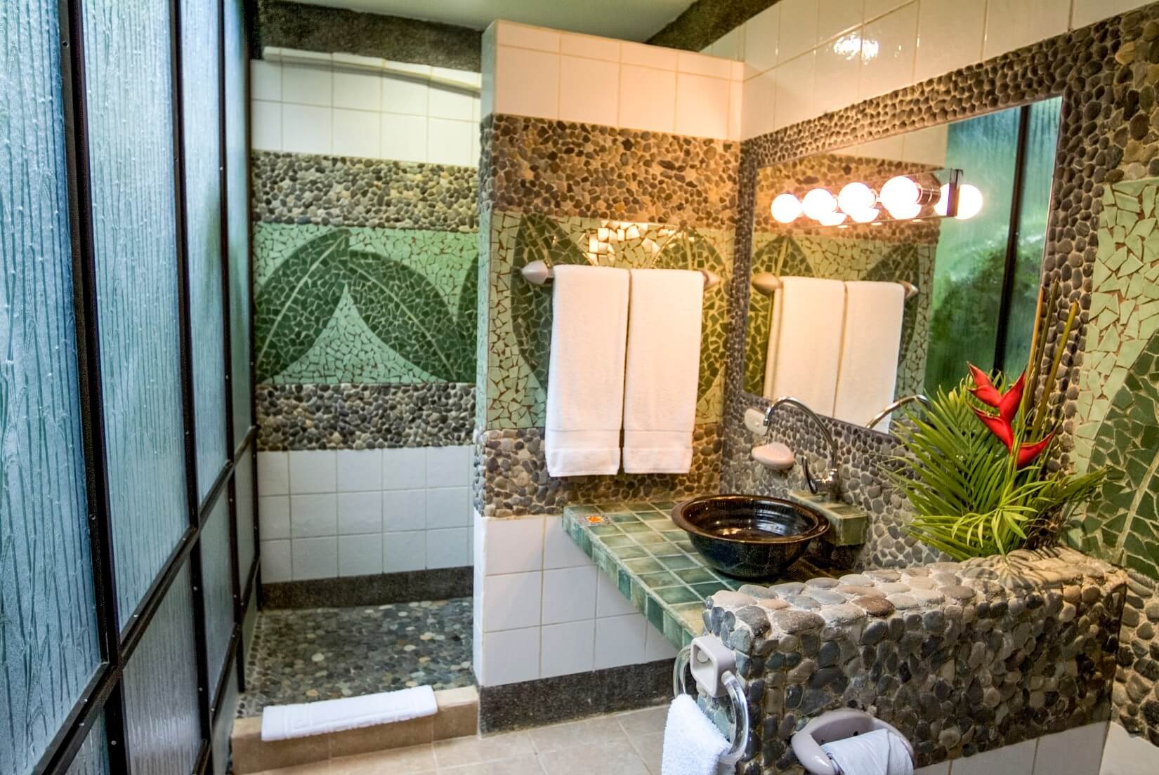 This bathroom features a walk-in shower and vanity area crafted with natural river stone and green mosaic leaf tiles, creating an earthy tropical aesthetic under diffused light from a textured glass panel.