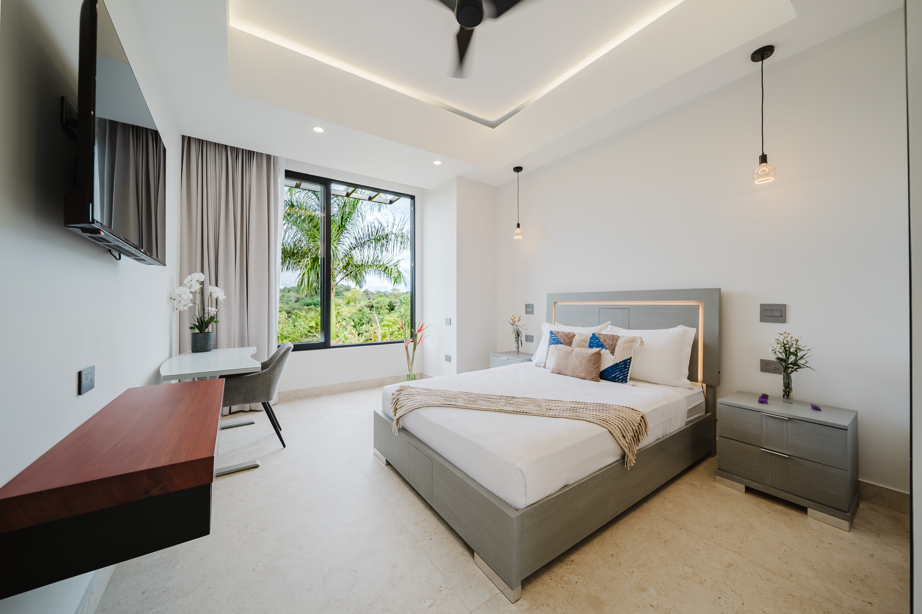A bright bedroom features a contemporary bed with integrated backlighting, a ceiling fan, and a large window presenting a verdant tropical view.