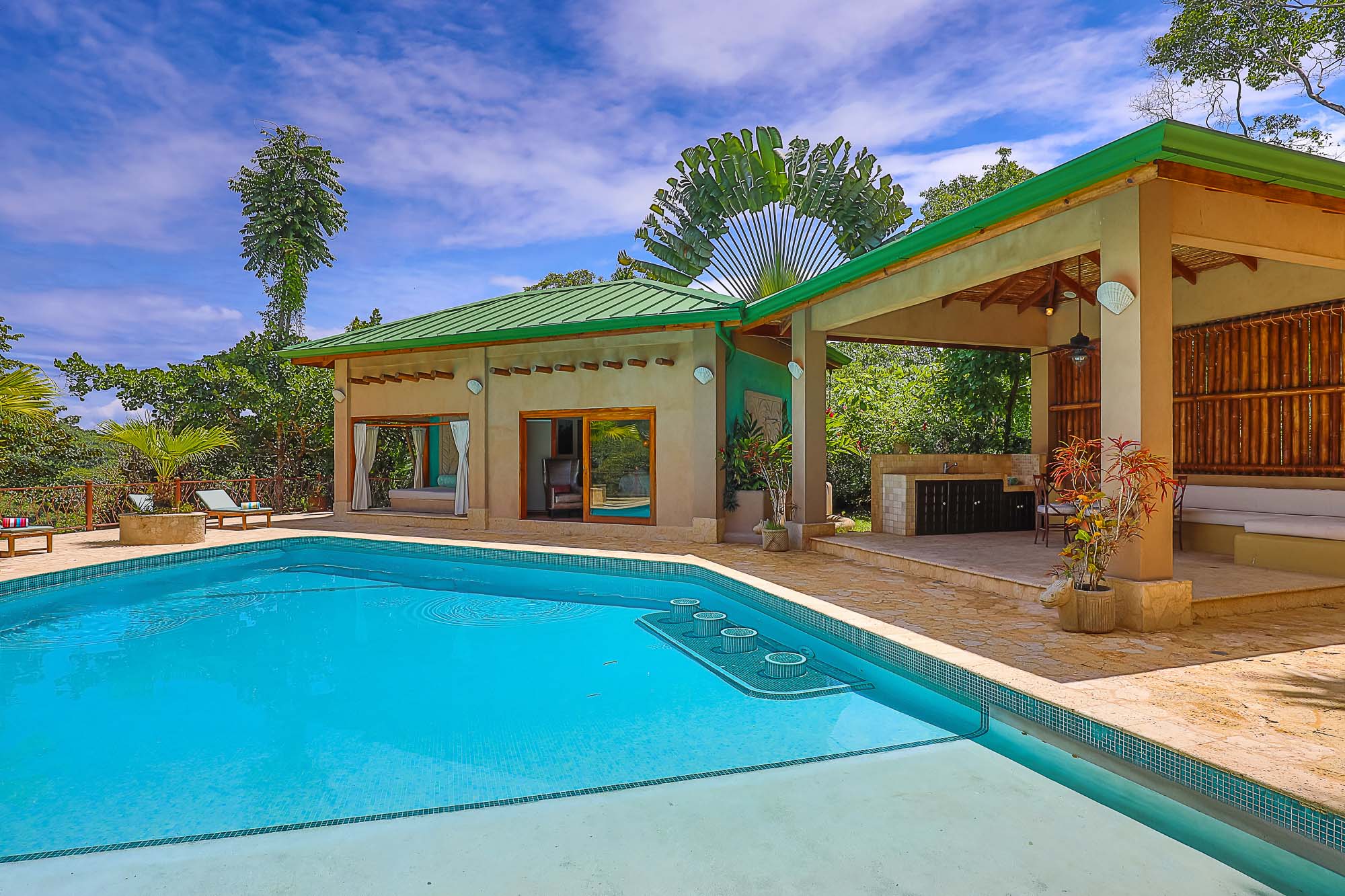 A bright blue swimming pool fronts a pool house with a visible bedroom and an open-air pavilion featuring an outdoor kitchen and lounge area, all surrounded by tropical greenery.