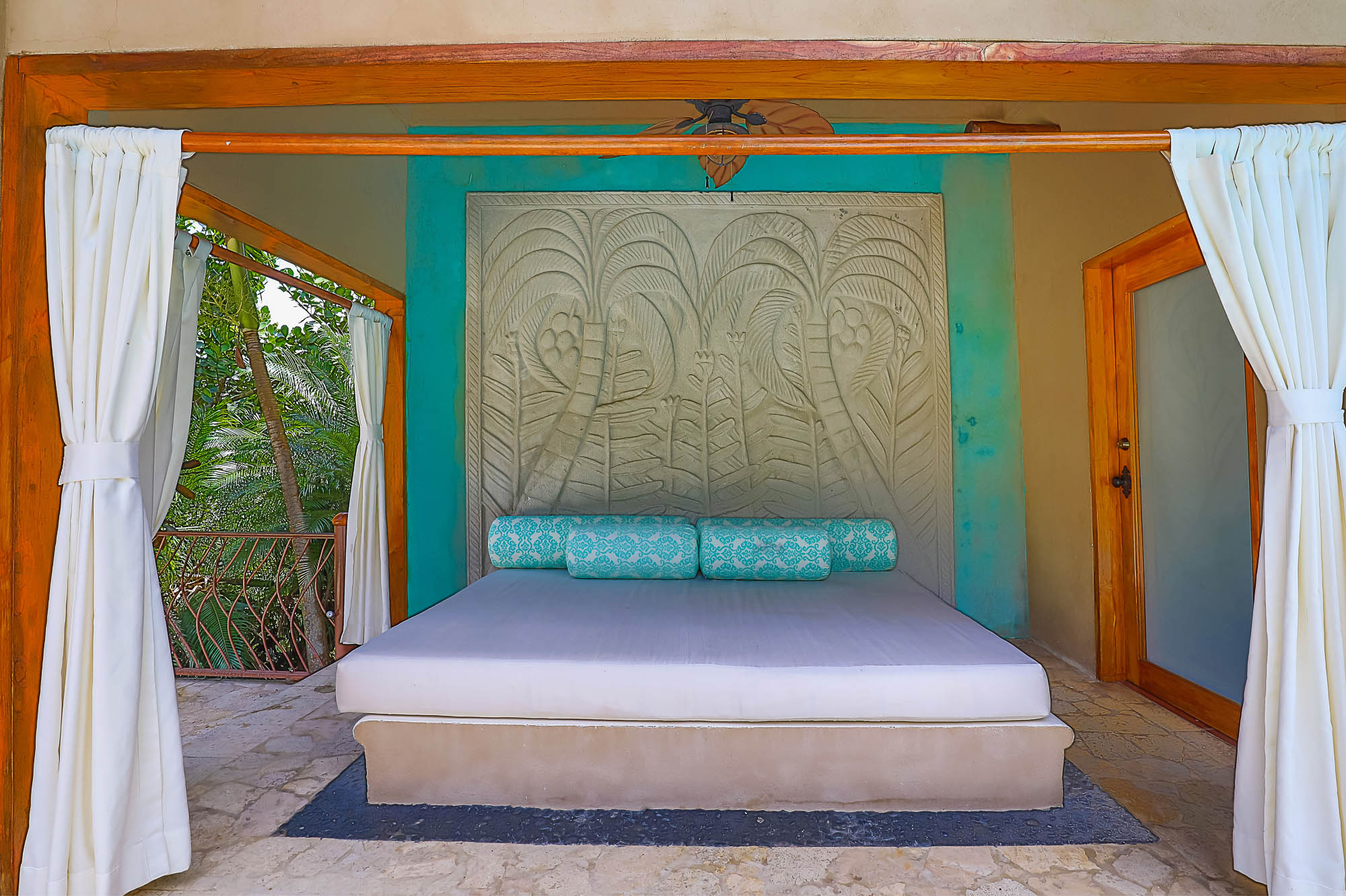 An open-air bedroom features a bed with a carved headboard and natural wood accents, overlooking a lush tropical rainforest.