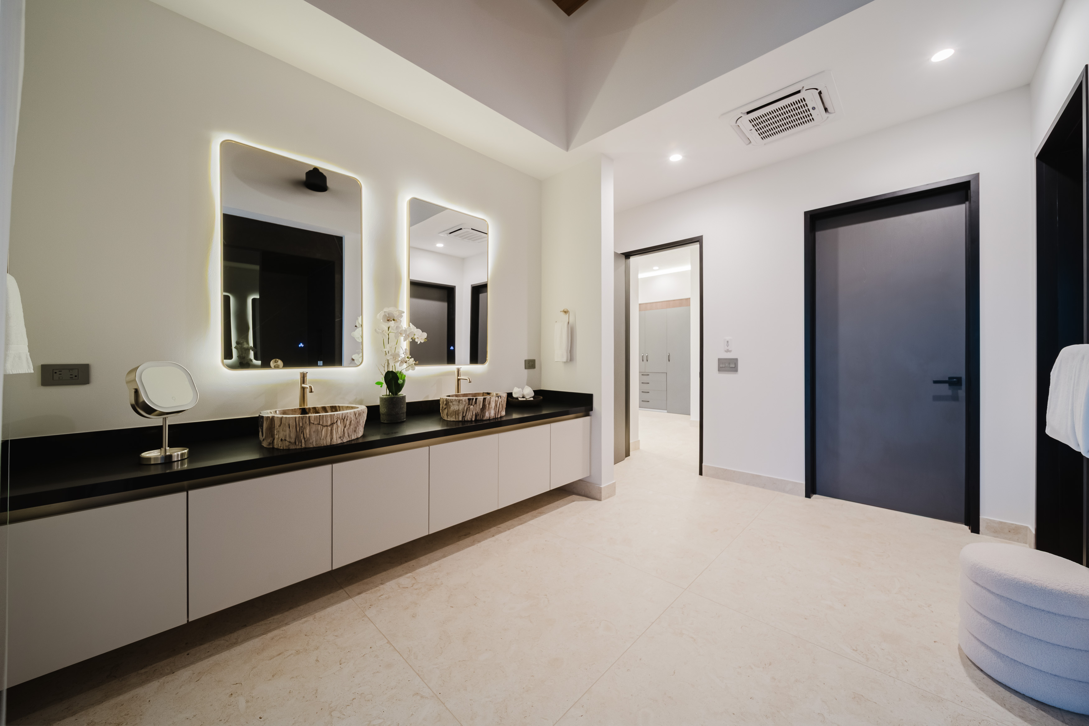 This spacious bathroom features a double vanity with natural stone vessel sinks, backlit mirrors, ceiling air conditioning, and flush doors leading to adjacent private areas.