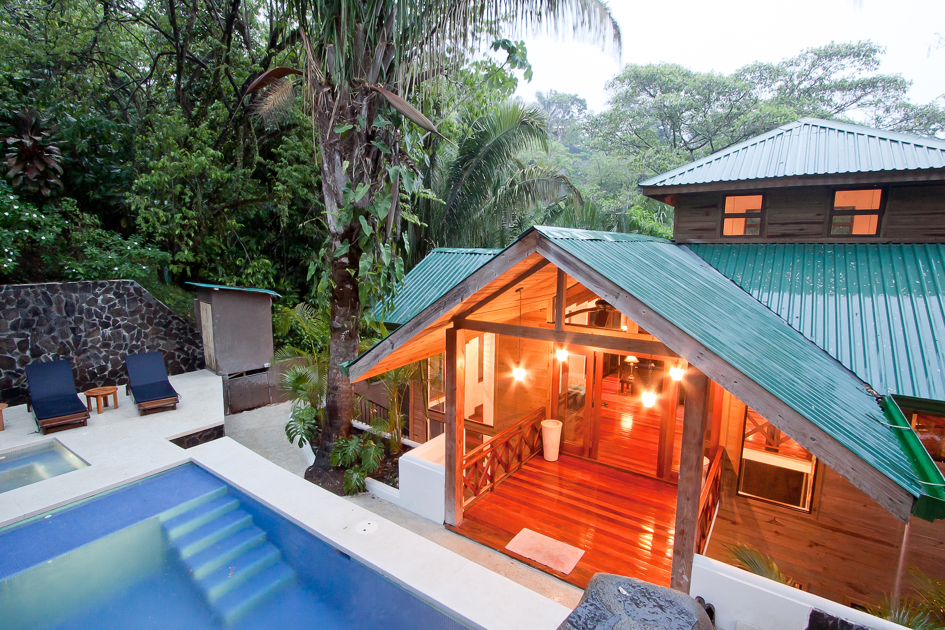 The multi-level wooden villa, featuring a green roof and warmly lit interior spaces, connects to a private pool and jacuzzi area, all immersed in a vibrant jungle environment.