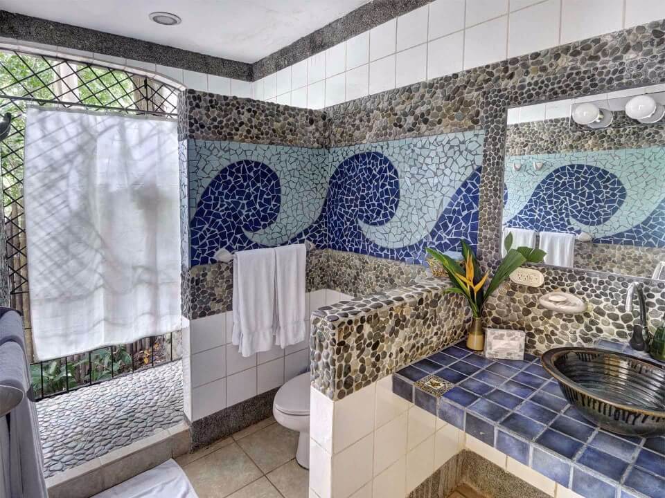 This bathroom features pebble walls and a blue mosaic wave pattern, a dark basin sink on a blue tiled vanity, and an open-air shower with a pebble floor that connects to the surrounding tropical greenery.