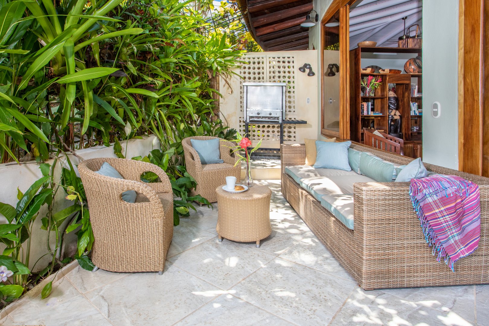 This outdoor terrace offers comfortable wicker lounge seating, bathed in dappled sunlight and surrounded by lush tropical foliage, with an open connection to the villa's interior.