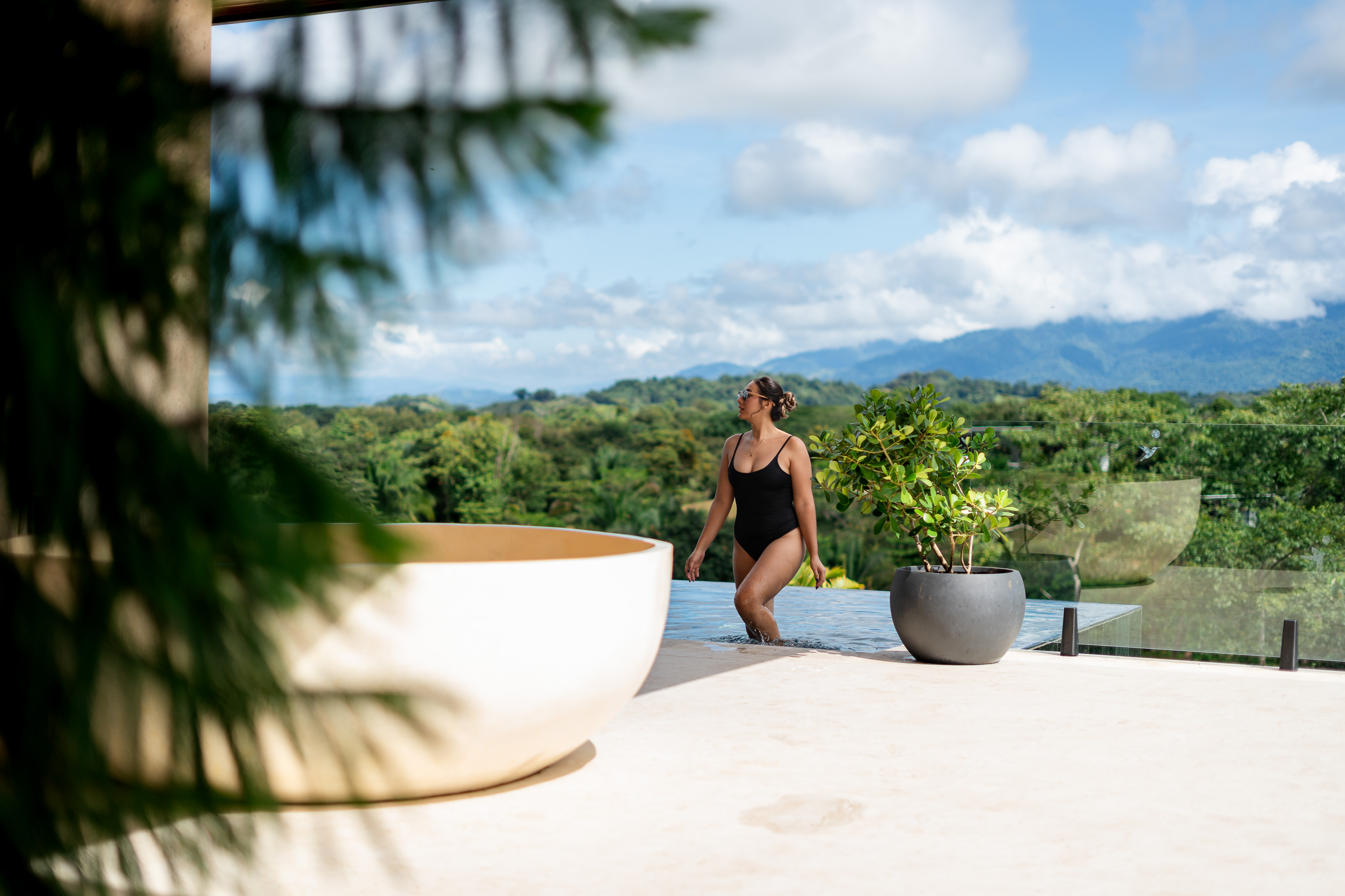 The outdoor infinity pool overlooks an expansive vista of tropical forests and distant mountains under a bright, cloudy sky.